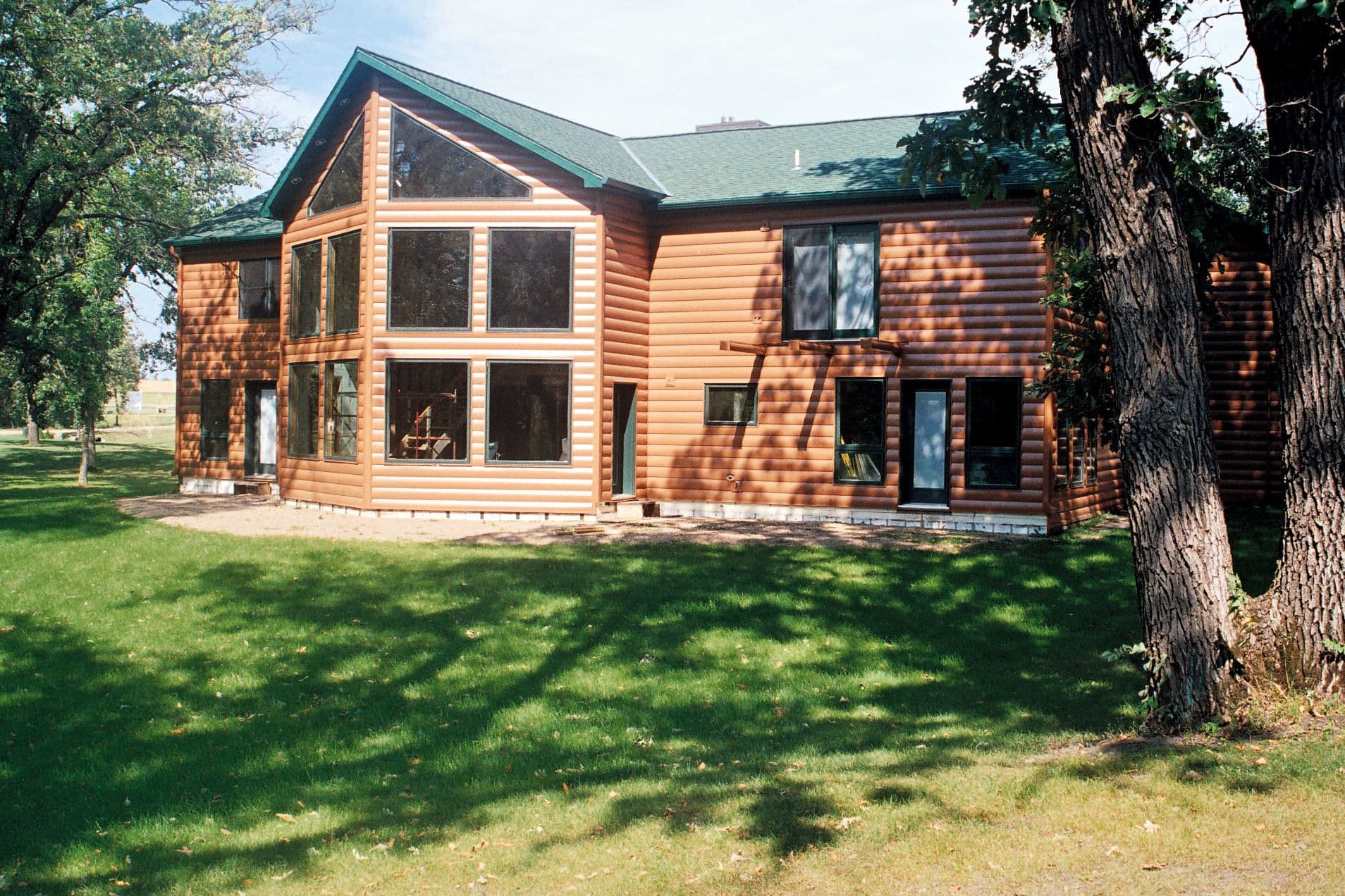 Two-story log cabin with green metal roof, large windows, and stone foundation, surrounded by mature trees and green lawn