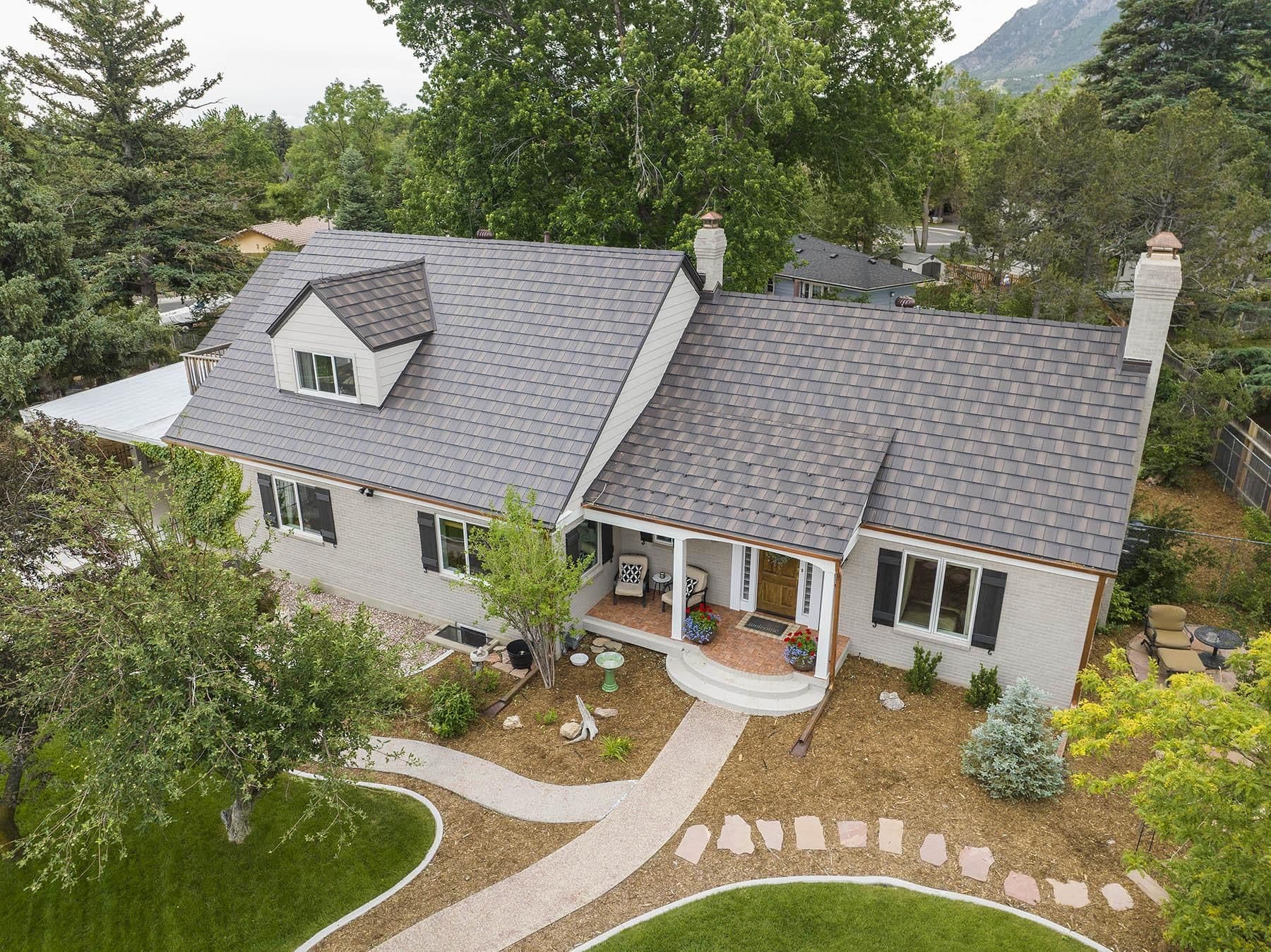 Aerial view of a modern white brick house with gray slate roof, dormer window, and brick patio surrounded by mature trees and landscaped yard with stepping stones