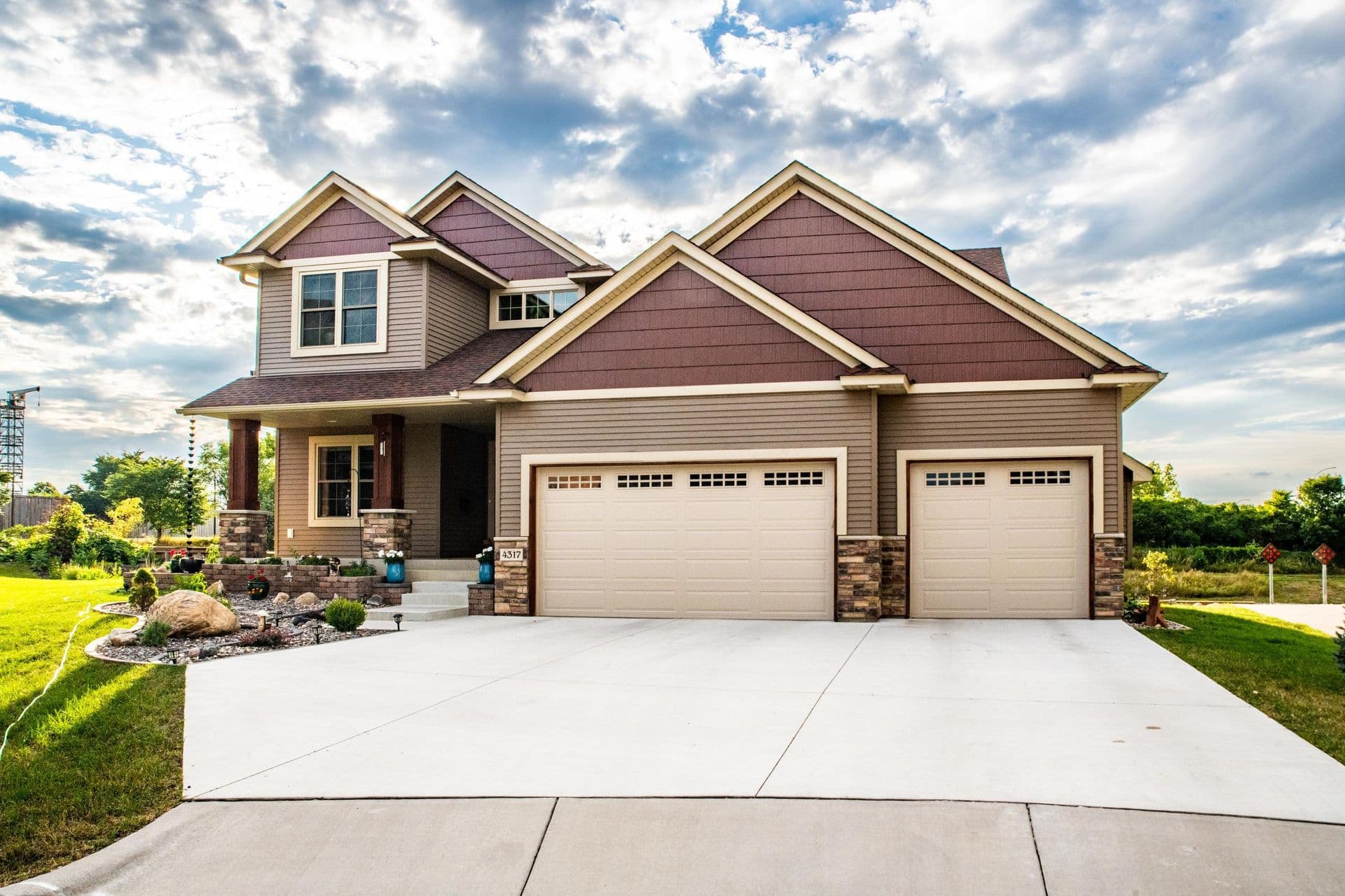 Two-story suburban home with tan siding, brown shingle roof, and two-car garage, featuring a front porch and landscaped yard with blue cistern