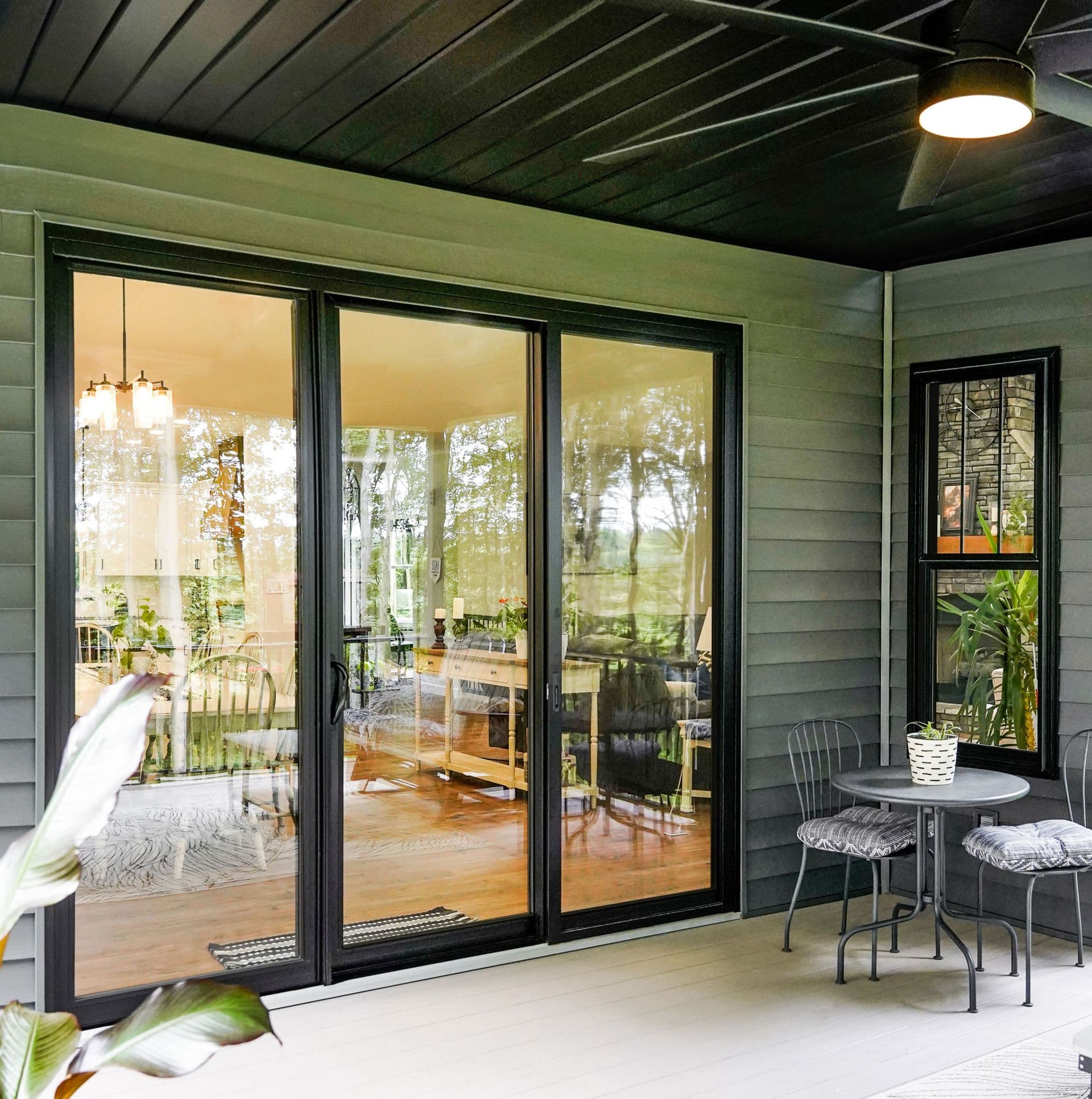 Modern screened porch with black-framed sliding glass doors opening to interior room with wood flooring, small bistro table and chairs, and potted plants