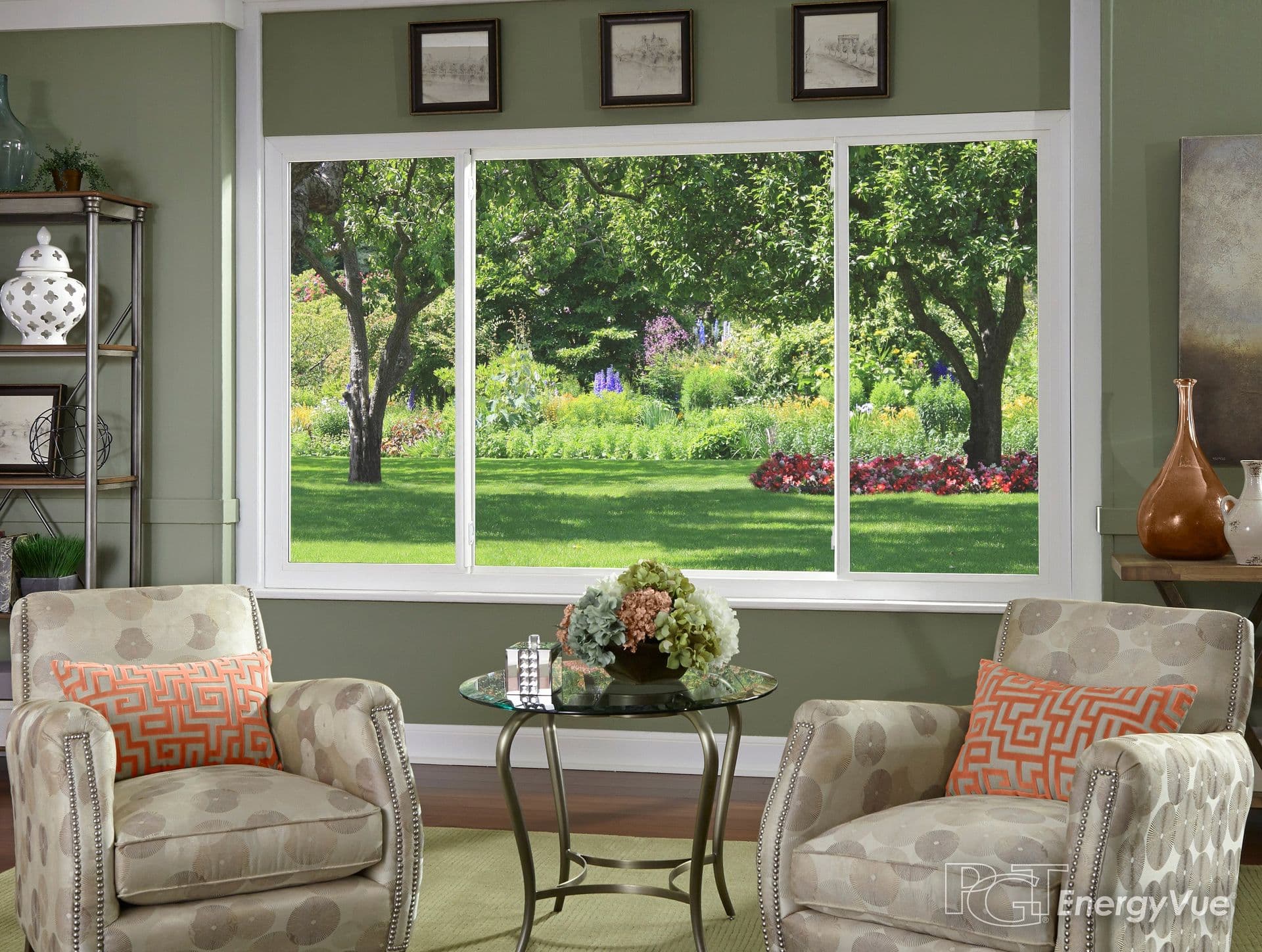 Living room with two upholstered armchairs facing a small round table, large triple-pane windows overlooking a garden with mature trees and flowering plants, sage green walls, and decorative shelving with accessories
