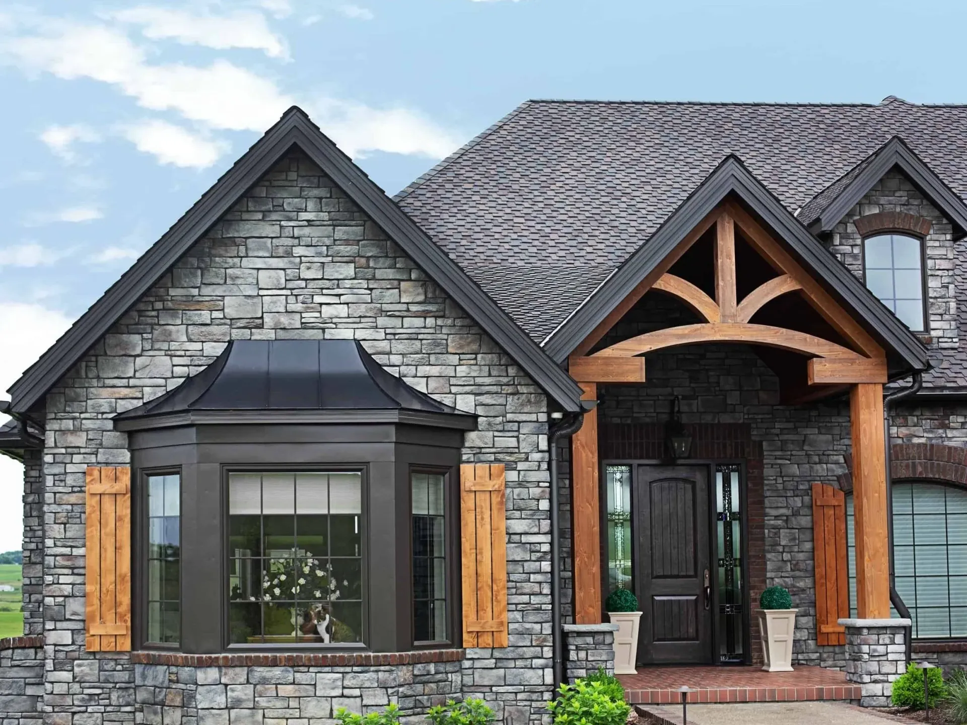 Contemporary stone home with peaked roof featuring a peaked bay window with dark shutters and wood trim on the left, arched wooden entry portico with black door on the right, and gray shingles above