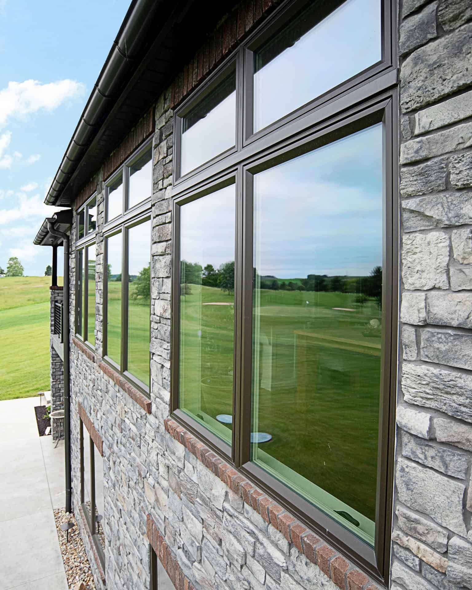 Row of large aluminum-framed windows on stone building overlooking rolling green fields and golf course with blue sky