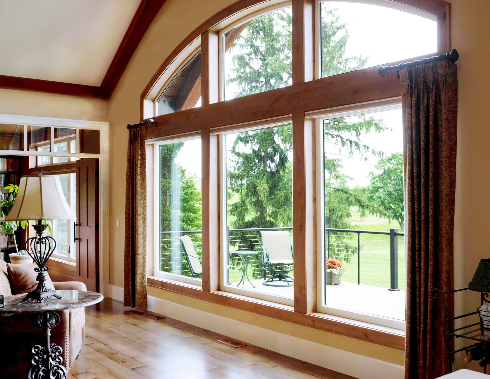 Sunlit living room with floor-to-ceiling windows and wood-trimmed glass doors overlooking a deck and forested landscape, burgundy curtains frame the view