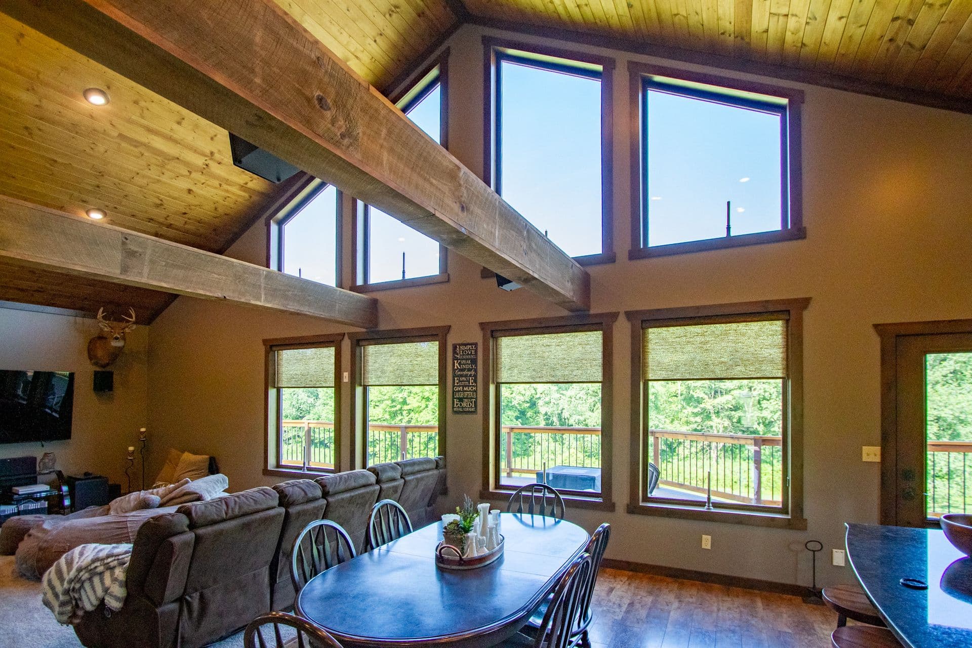 Spacious living room with vaulted wooden ceiling, exposed beams, large windows overlooking forested landscape, gray sectional sofa, blue dining table with chairs, and deer mount on wall