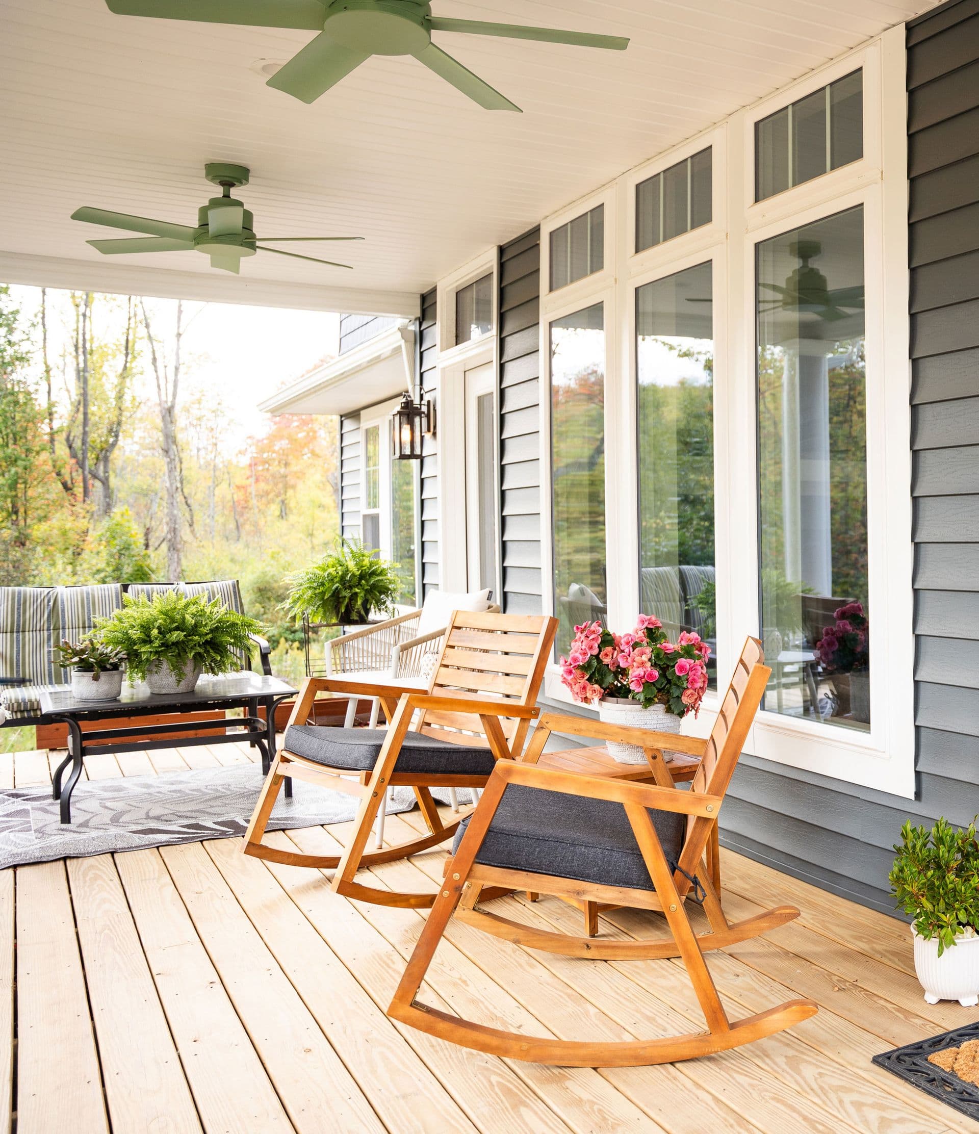 Covered porch with wooden rocking chairs, pink potted flowers, ceiling fans, and expansive windows overlooking autumn foliage