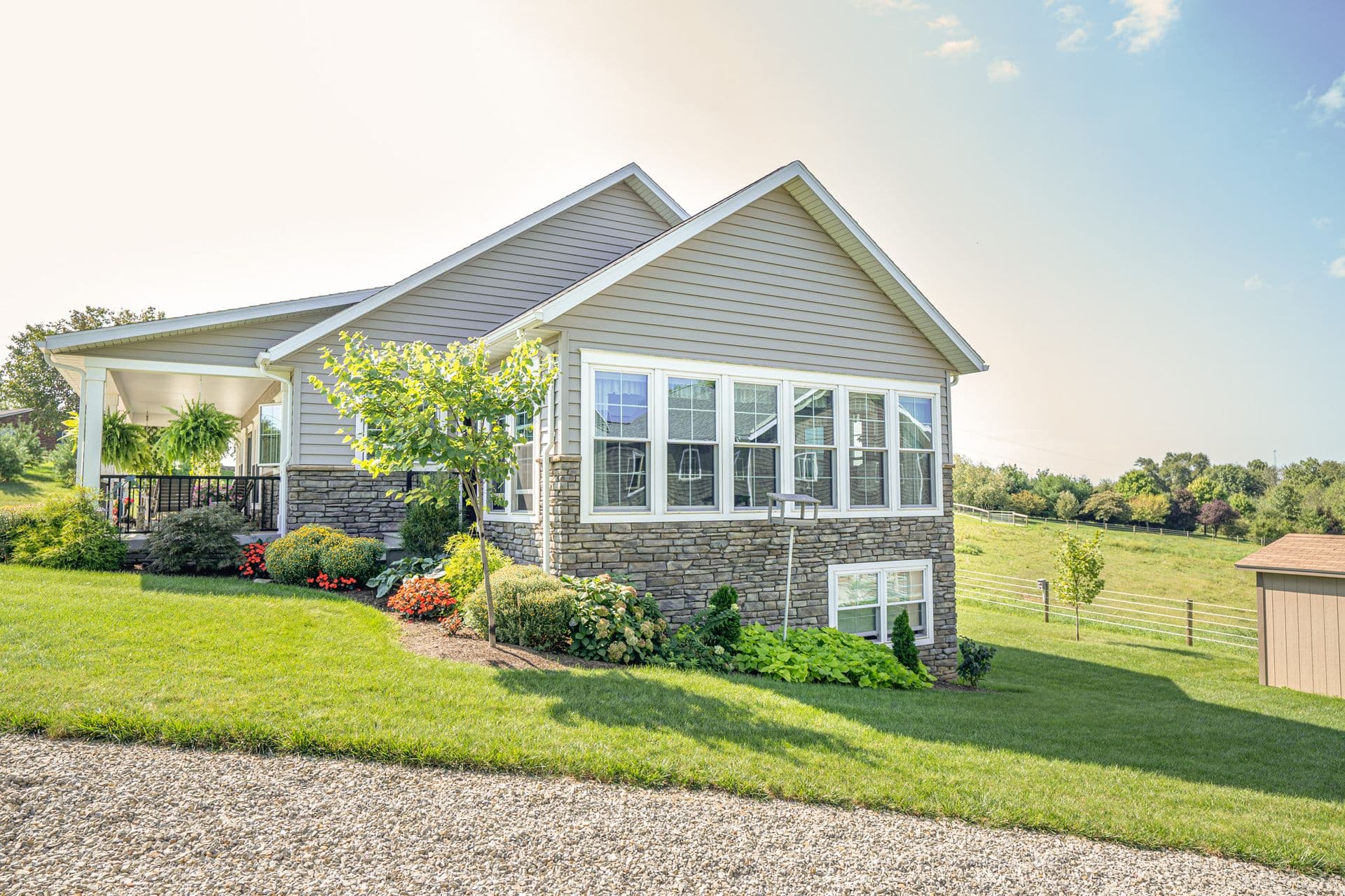 Provia - Modern single-story home with stone and siding exterior, covered porch with railing, multiple large windows, landscaped front yard with flowering plants and shrubs, situated on rural acreage with pasture fencing visible