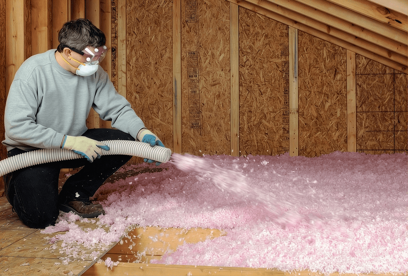 Worker in safety goggles and mask blowing pink fiberglass insulation into attic space between wooden wall studs and OSB sheathing
