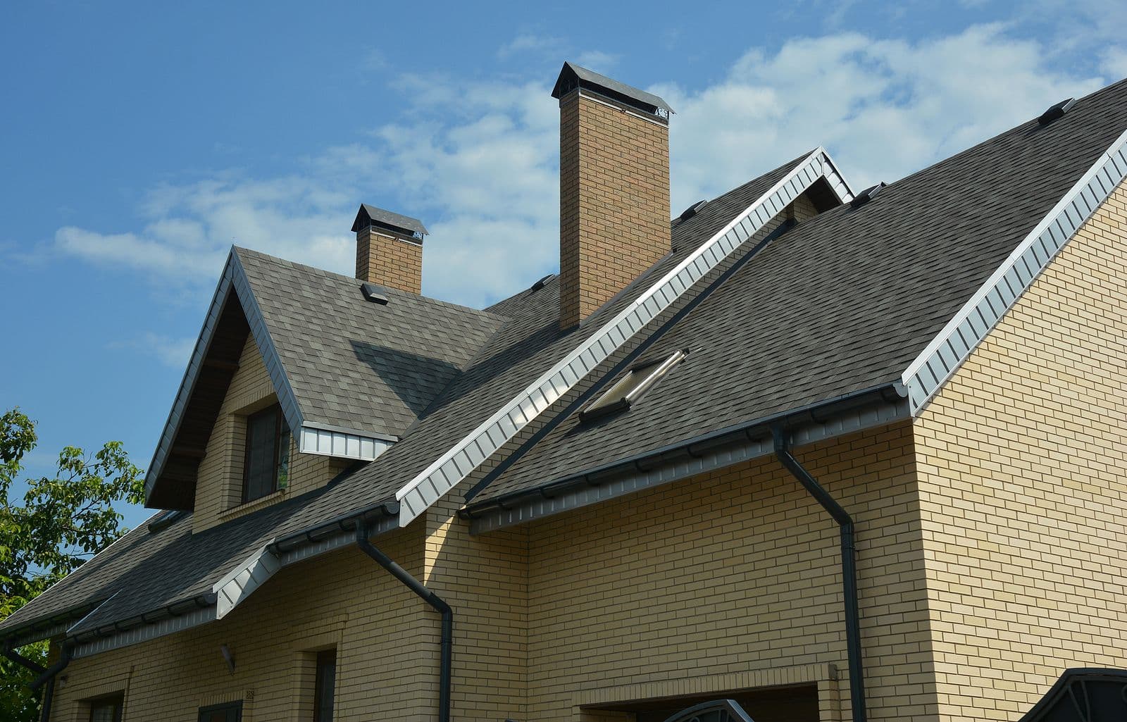 Brick residential house with steep pitched roof, two tall chimneys, white soffit and fascia trim, and gabled dormer windows against a blue sky