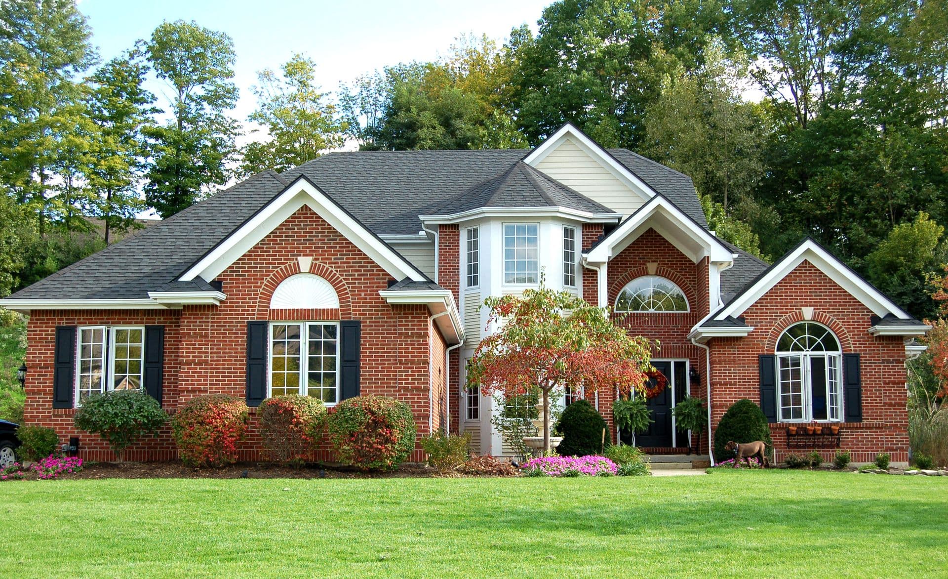 Two-story red brick house with white trim, multiple gabled roofs, arched windows, black shutters, manicured lawn, flowering shrubs, and mature trees in background