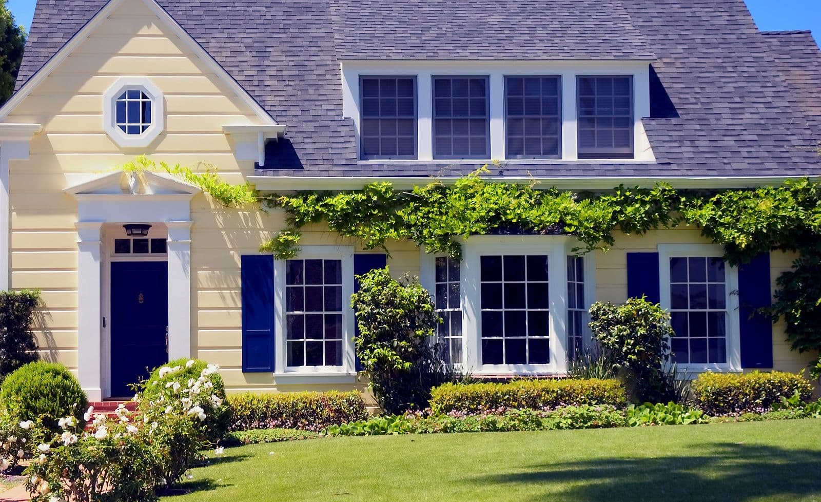 Cream-colored house with purple roof and dark blue shutters, featuring a circular window and covered entry. Ivy-covered facade with four large windows, manicured front lawn and flowering shrubs