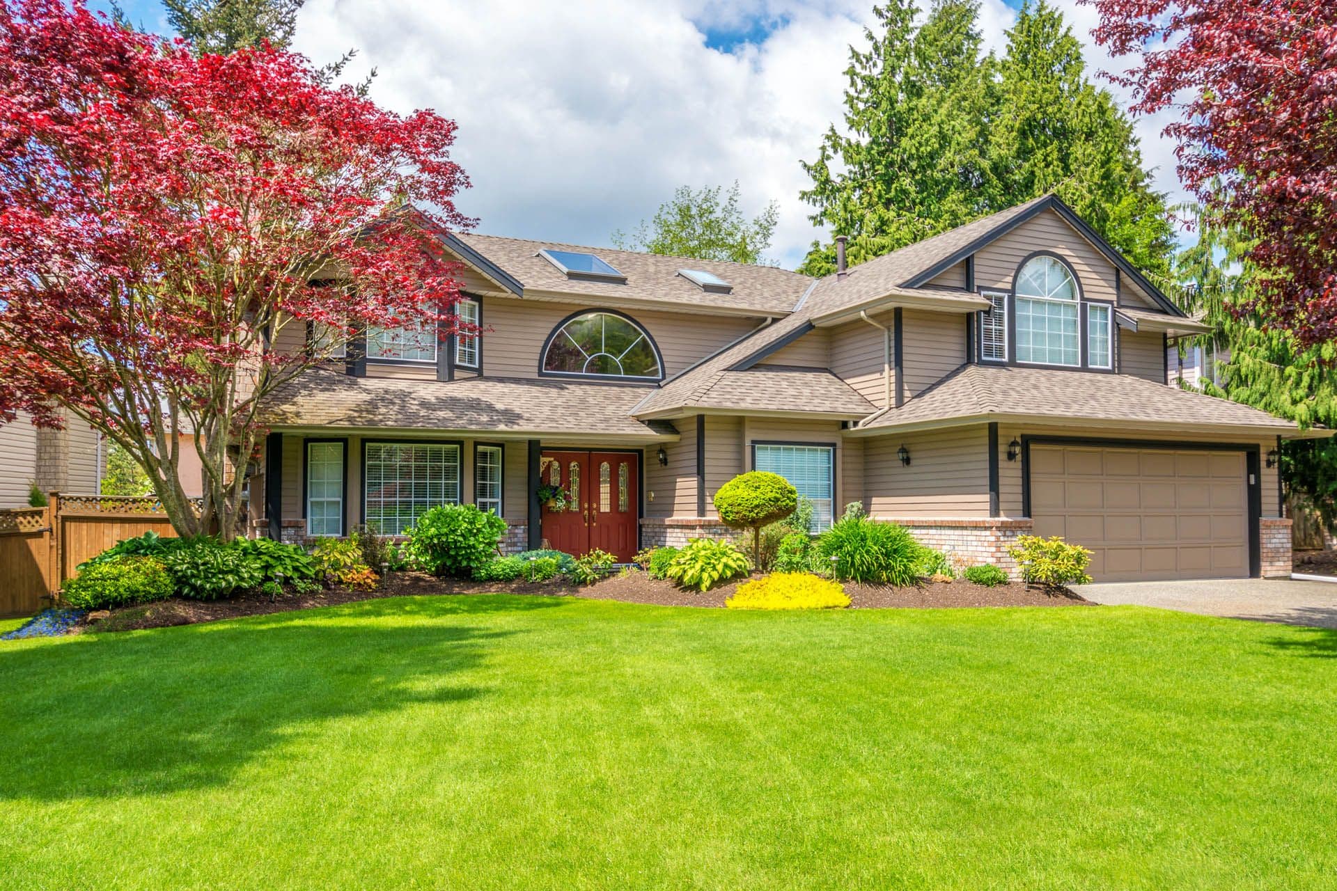 Two-story suburban home with beige siding, two-car garage, and burgundy front door, surrounded by manicured landscaping including red Japanese maple trees and green shrubs, with well-maintained lawn