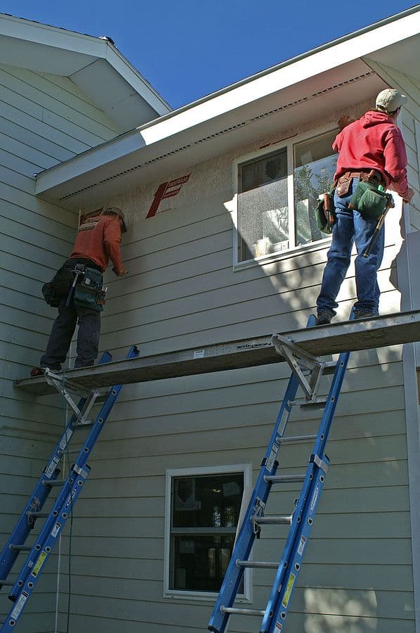 Two construction workers on scaffolding installing or repairing siding on a light gray house exterior