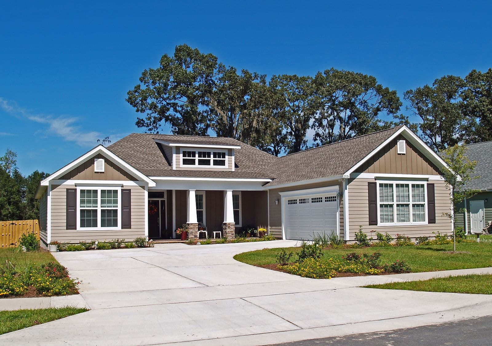 Modern single-story home with beige siding, two-car garage, covered porch with columns, and dormer window, surrounded by mature oak trees and landscaped front yard