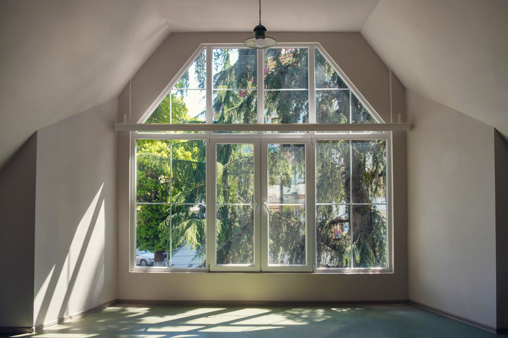 Bright attic room with large multi-pane windows overlooking tall evergreen trees, pendant light fixture, and sunlight streaming across teal floor