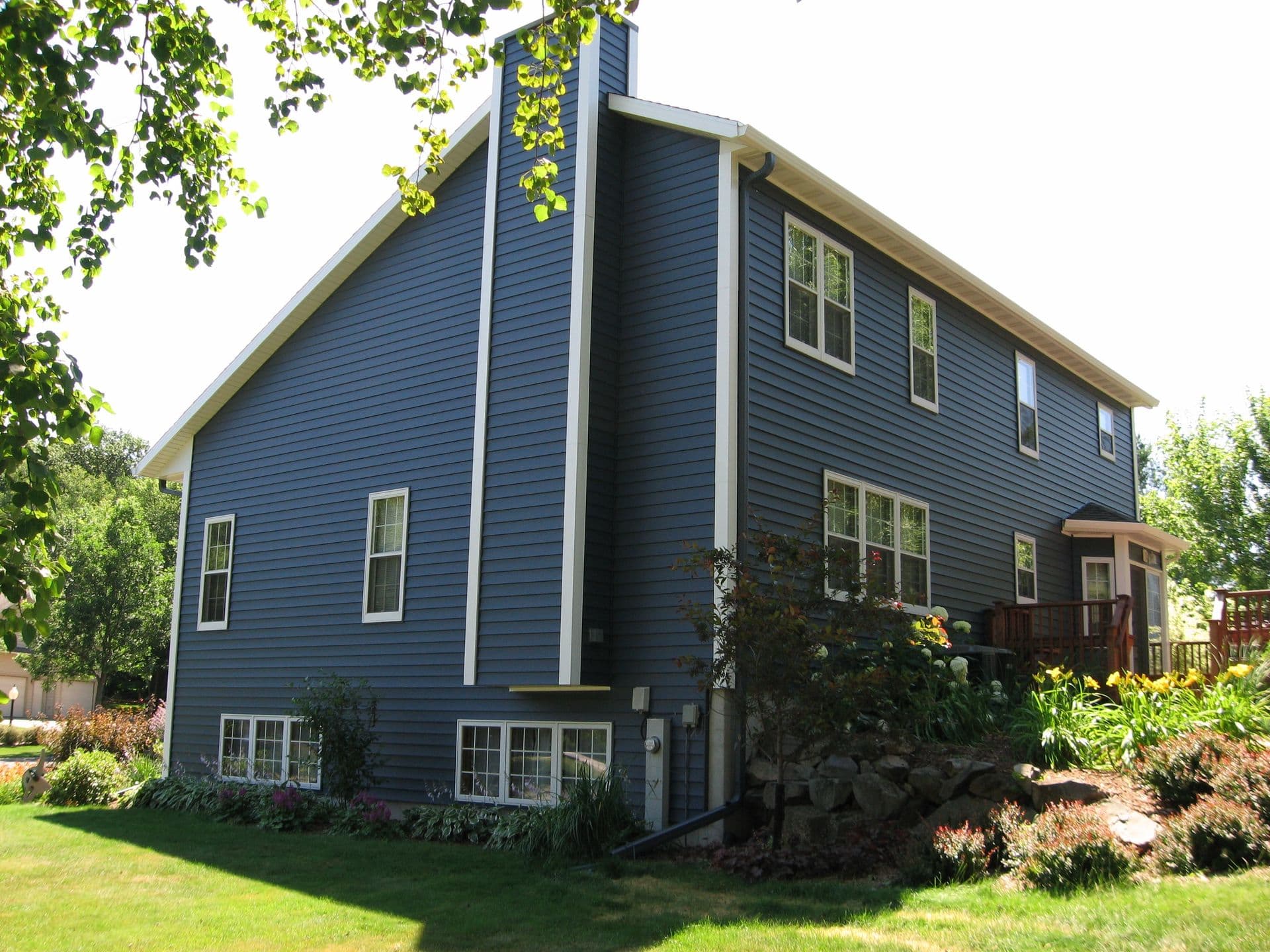 Dark blue modern farmhouse with elevated foundation, multiple windows, wooden deck, surrounded by mature trees and landscaped gardens