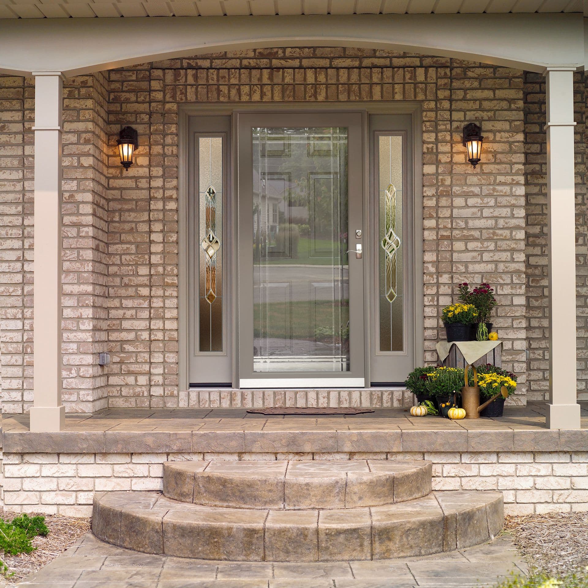 Front porch entry with taupe double doors flanked by sidelights, brick facade, curved stone step, lantern sconces, and fall mums and pumpkins on display
