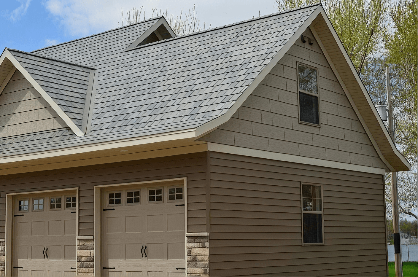 Modern residential home with gray metal roof, beige siding, two-car garage with paneled doors, and gabled dormers