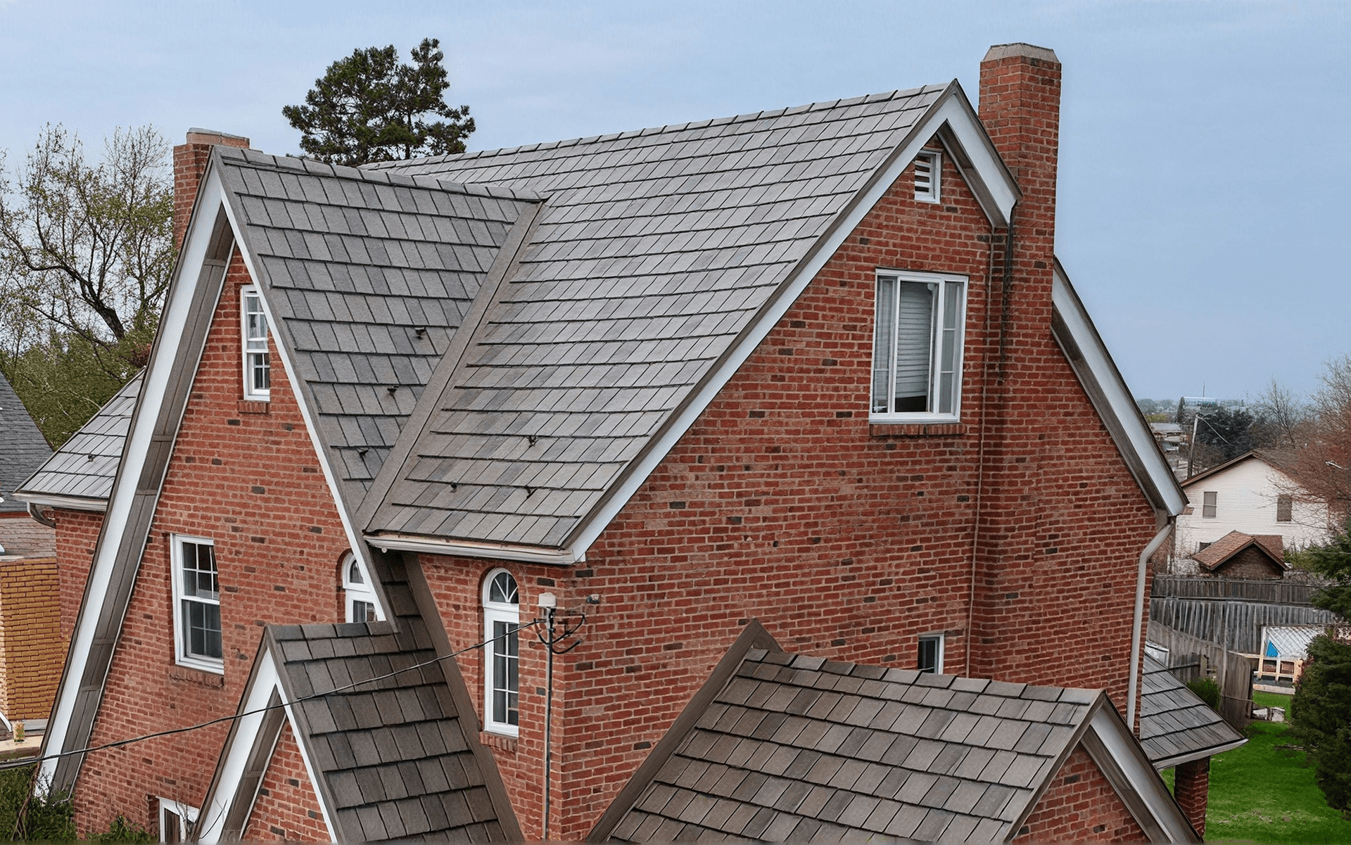Red brick house with multiple gray metal-shingle roofs, white trim, and tall brick chimney, showcasing residential roofing styles and architectural design