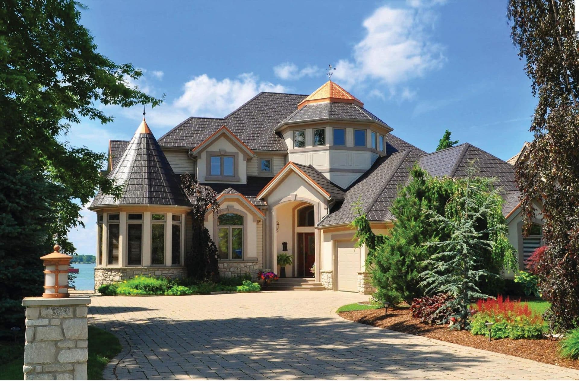 Waterfront luxury home with turret and curved tower features, stone and stucco exterior, copper accents on peaked roofs, surrounded by manicured landscaping and ivy-covered trees