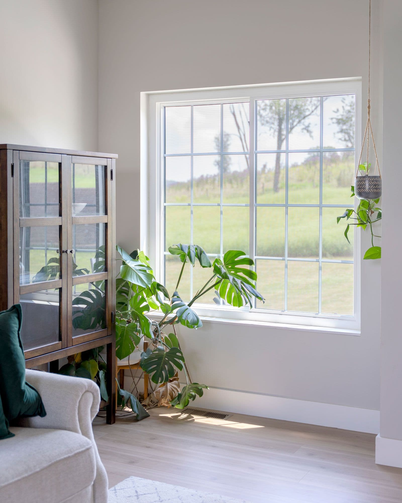 Bright living room with large gridded windows overlooking green fields, featuring potted plants including a monstera and hanging succulent, cream upholstered chair, and wooden glass cabinet