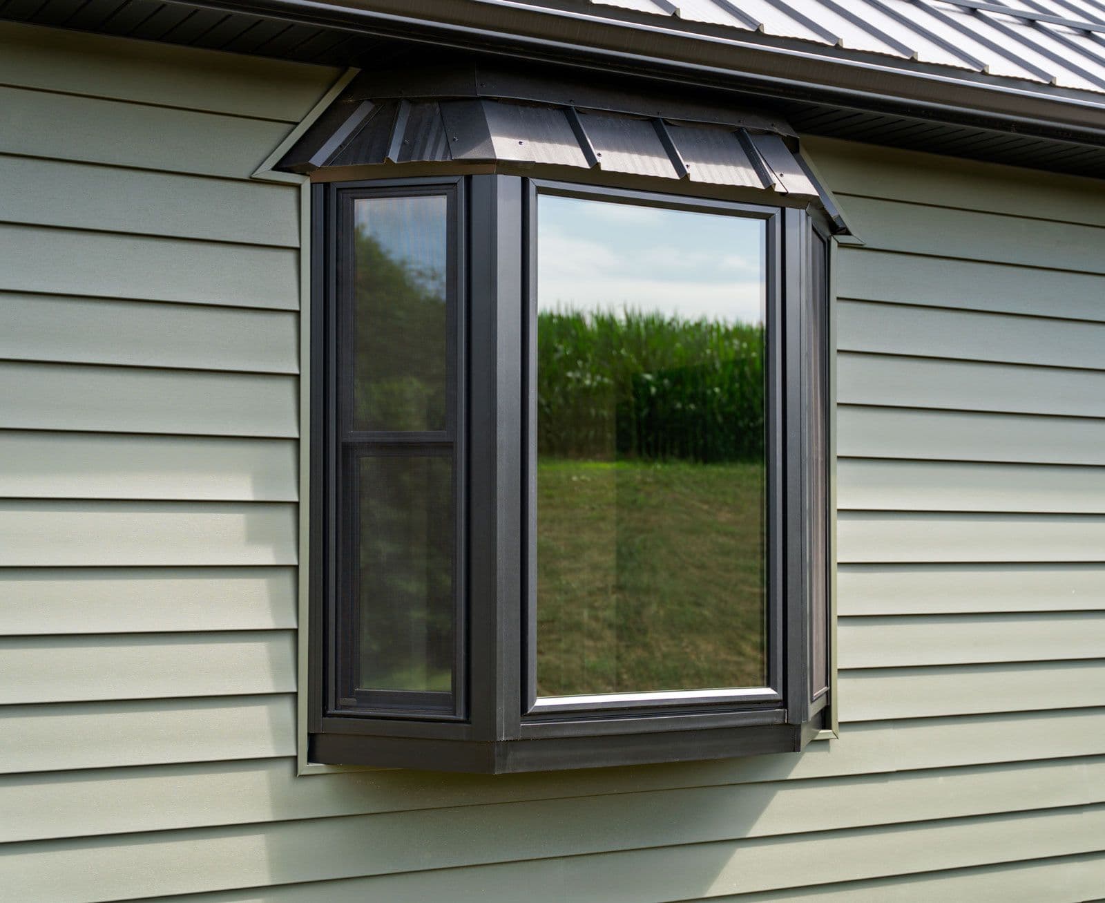 Modern black garden window with casement door and glass panes projecting from beige house siding, overlooking rural field and cornstalk backdrop