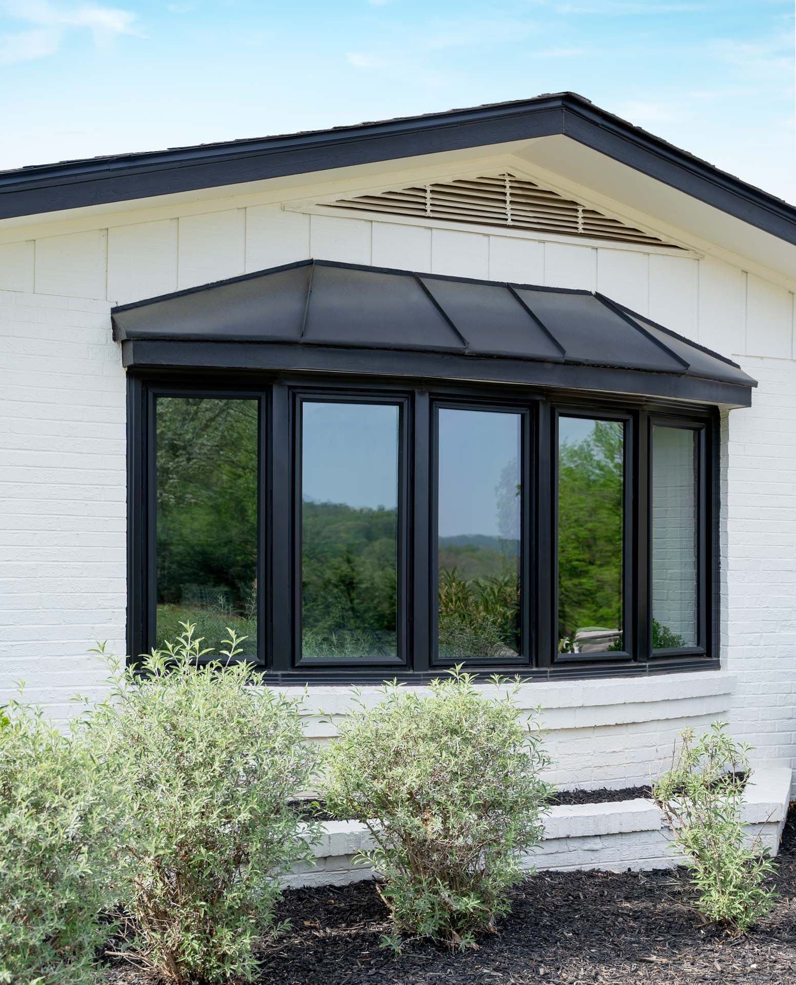 Modern white house with black-framed multi-panel windows and sloped awning, overlooking green landscape with shrubs in foreground