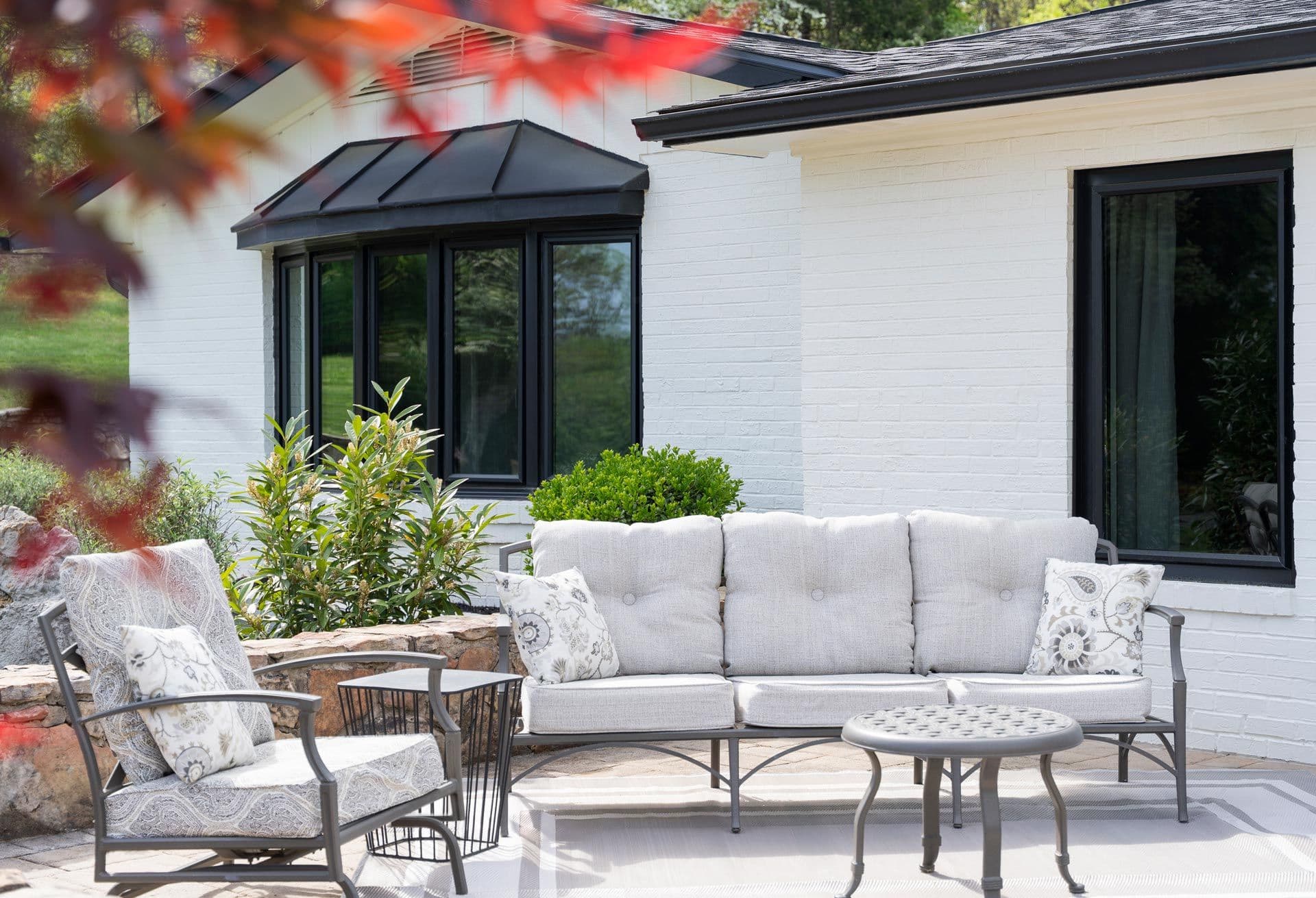 Modern patio with beige upholstered sofa, armchair, and metal coffee table in front of white brick house with black-framed windows and glass door