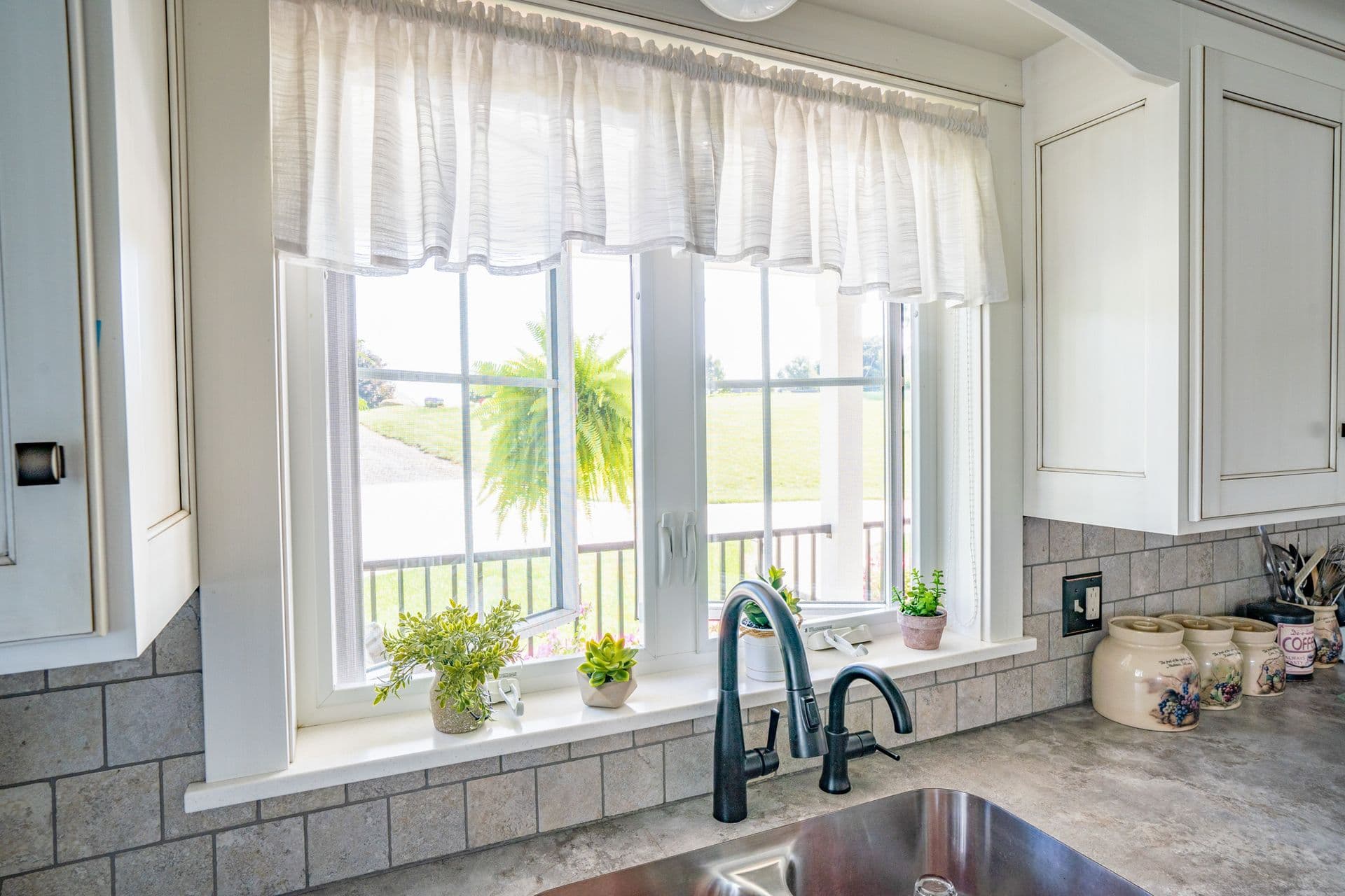 Kitchen sink with black faucet below window with sheer curtains, potted plants on sill, and view of landscaped yard with palm tree