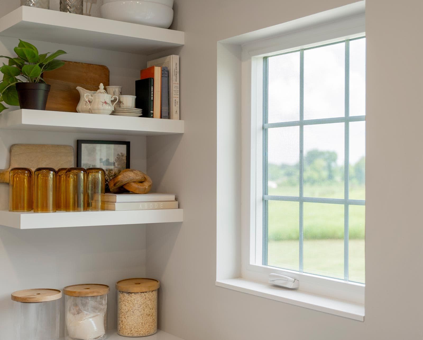 Modern kitchen shelving with decorative and functional items including books, potted plant, dishware, amber glass jars, and wooden storage containers, beside a large window overlooking green landscape