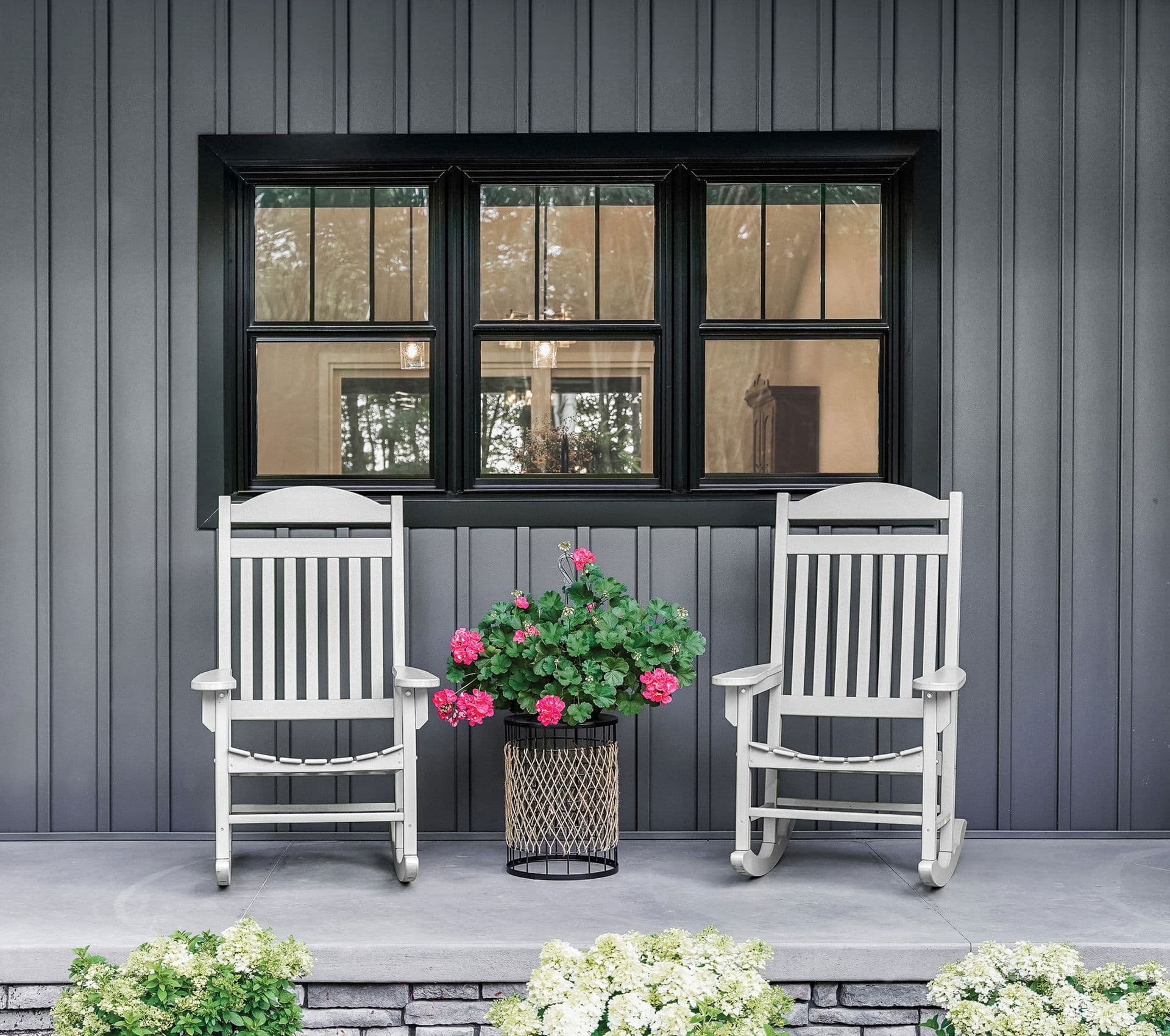 Two white rocking chairs flank a planter with pink flowers on a gray porch, in front of a black-framed window on a corrugated metal wall
