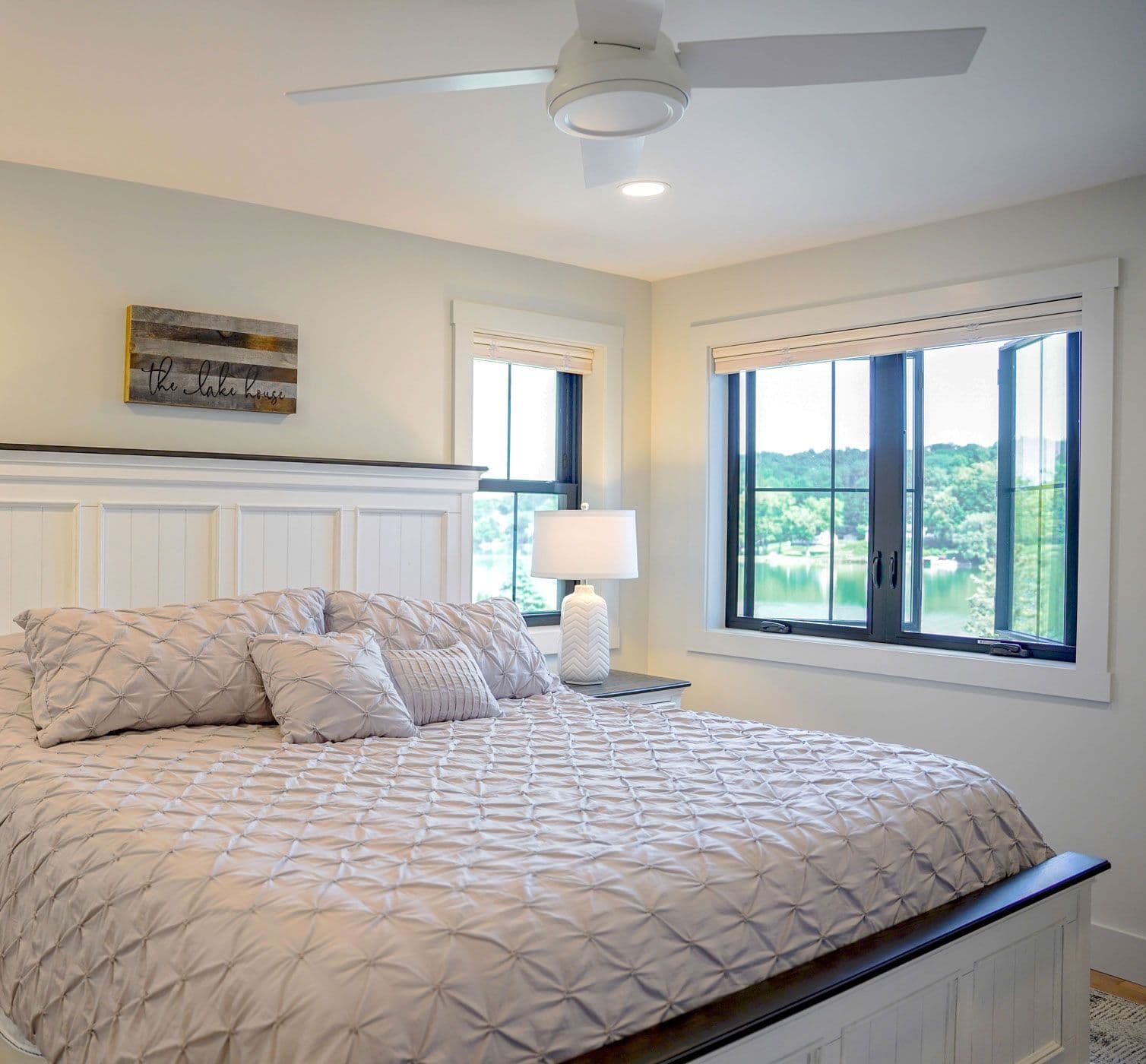Modern lakeside bedroom with white shiplap headboard, quilted bedding, ceiling fan, and expansive windows overlooking forested shoreline