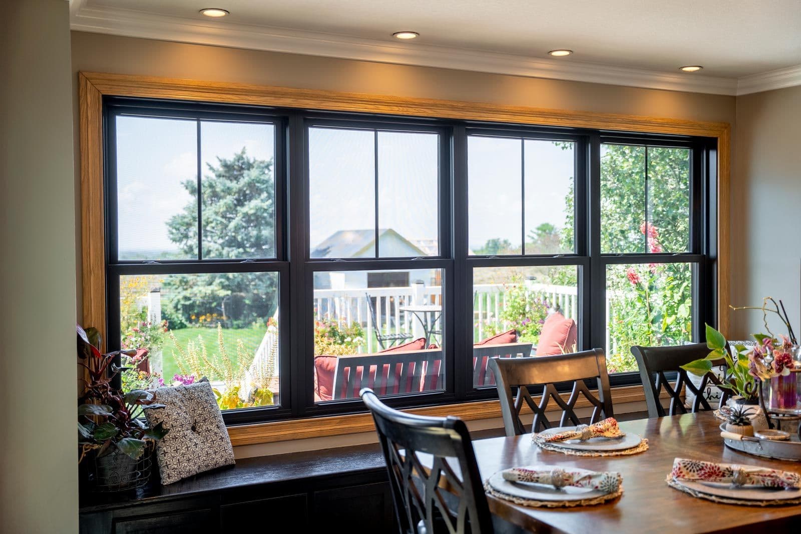 Dining room with large black-framed windows overlooking a garden and deck, wooden table with place settings, and potted plants on the windowsill