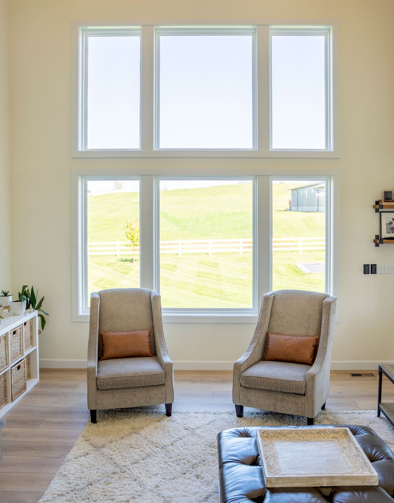 Modern living room with two beige armchairs facing large windows overlooking a rural landscape with white fencing and green fields