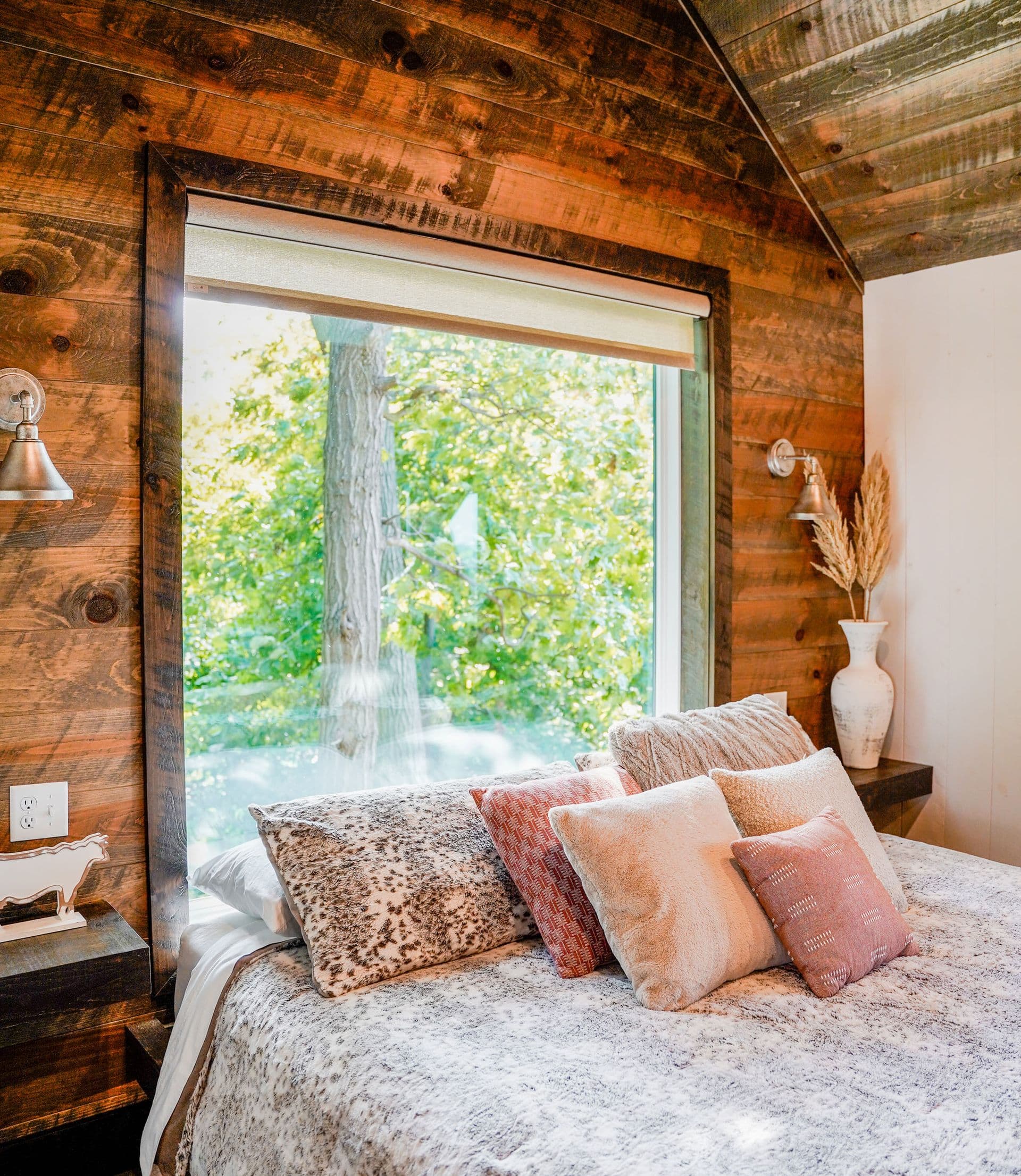 Rustic log cabin bedroom with large sliding glass door overlooking trees, bed with decorative pillows, exposed wood beams, and bedside lighting