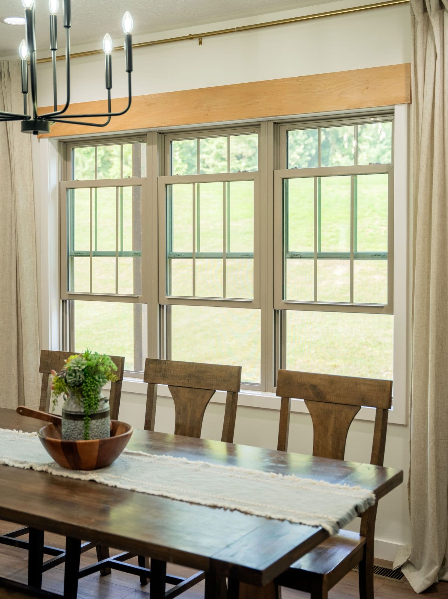 Modern dining room with wooden table, chairs, and potted plant in front of large multi-pane windows overlooking green landscape, lit by contemporary metal chandelier