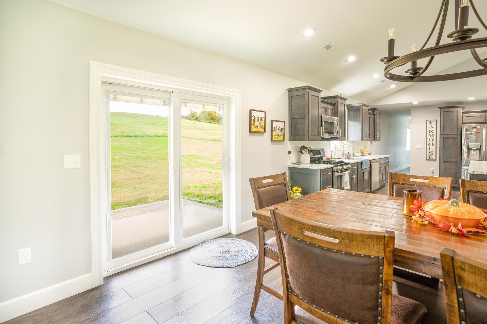 Open-concept kitchen and dining room with wooden table, brown leather chairs, gray cabinetry, and French doors opening to a rural pasture view