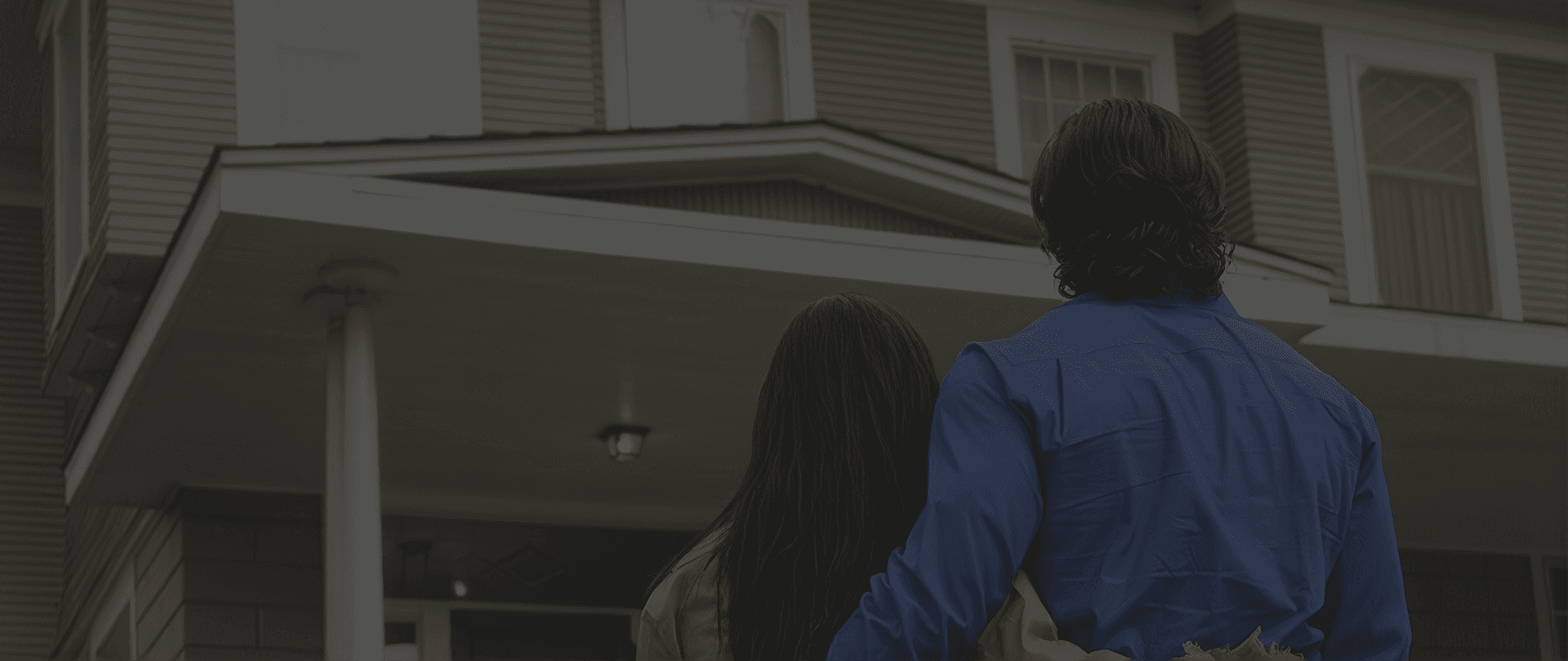 Couple standing together looking up at a two-story brick house with a second-floor dormer and front porch