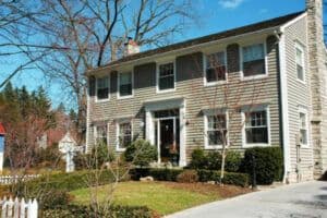 Two-story colonial-style house with beige siding, black shutters, and front porch, surrounded by landscaped grounds with bare trees and evergreens