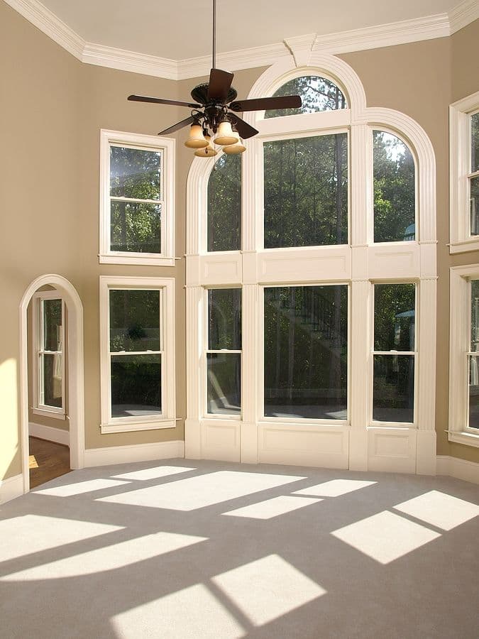 Sunlit room with cream walls and ceiling fan, featuring multiple tall windows with white trim and arched transom windows overlooking green trees