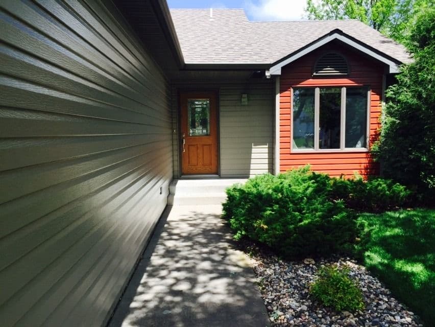 Single-story house exterior with red-orange siding, wooden front door, large window, and attached garage with green siding, surrounded by green shrubs and gravel landscaping