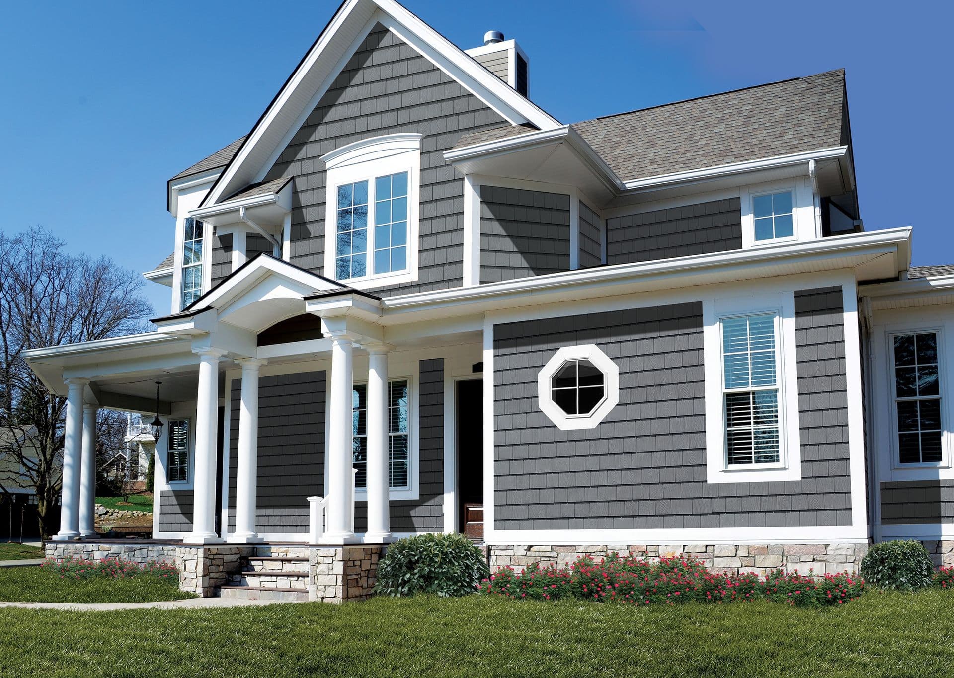 Contemporary two-story home with gray shingle siding, white trim, front porch with columns, stone foundation, and manicured lawn under clear blue sky
