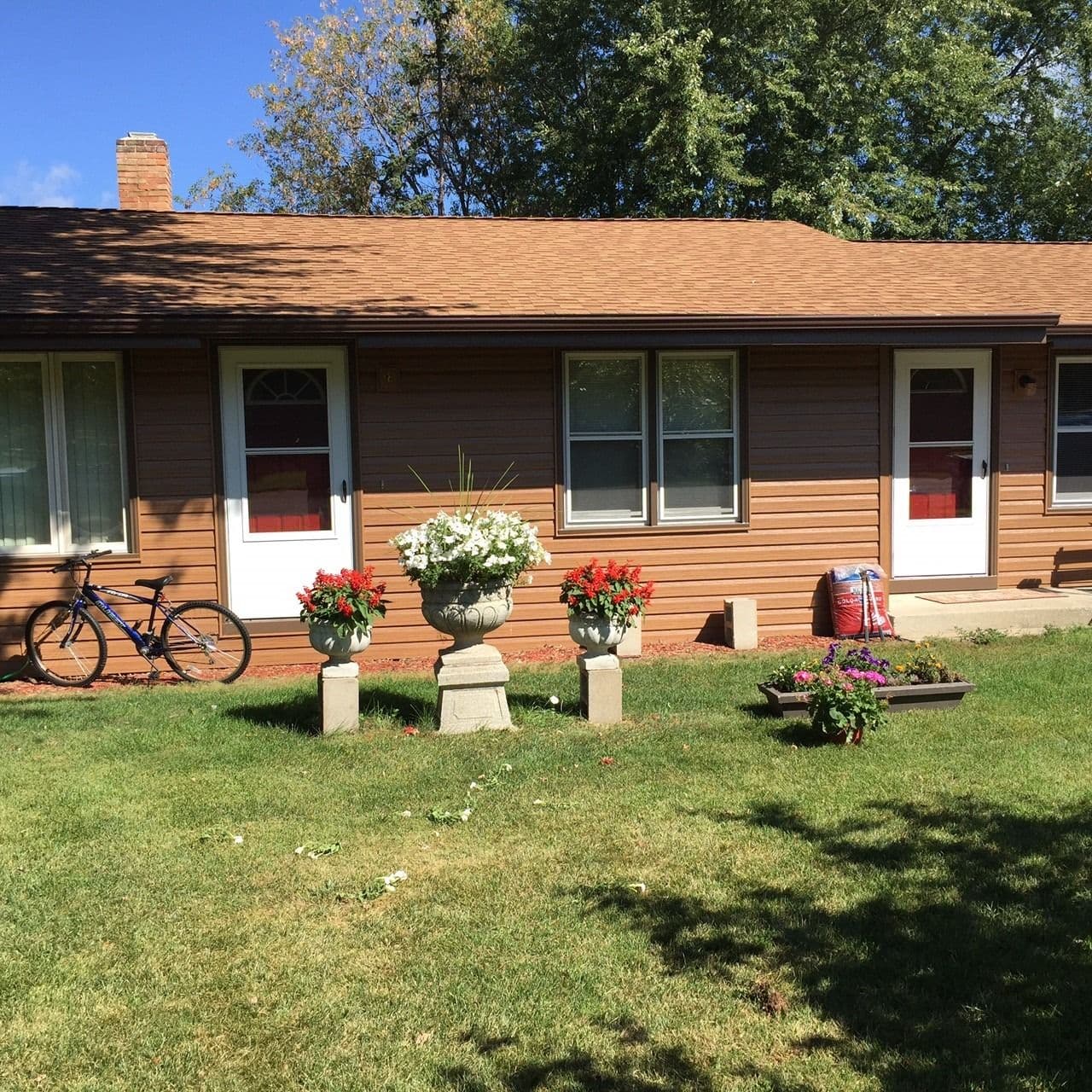 Single-story ranch house with wood siding, red brick chimney, and multiple windows, surrounded by landscaped front lawn with potted red and white flowers and a bicycle parked on the left