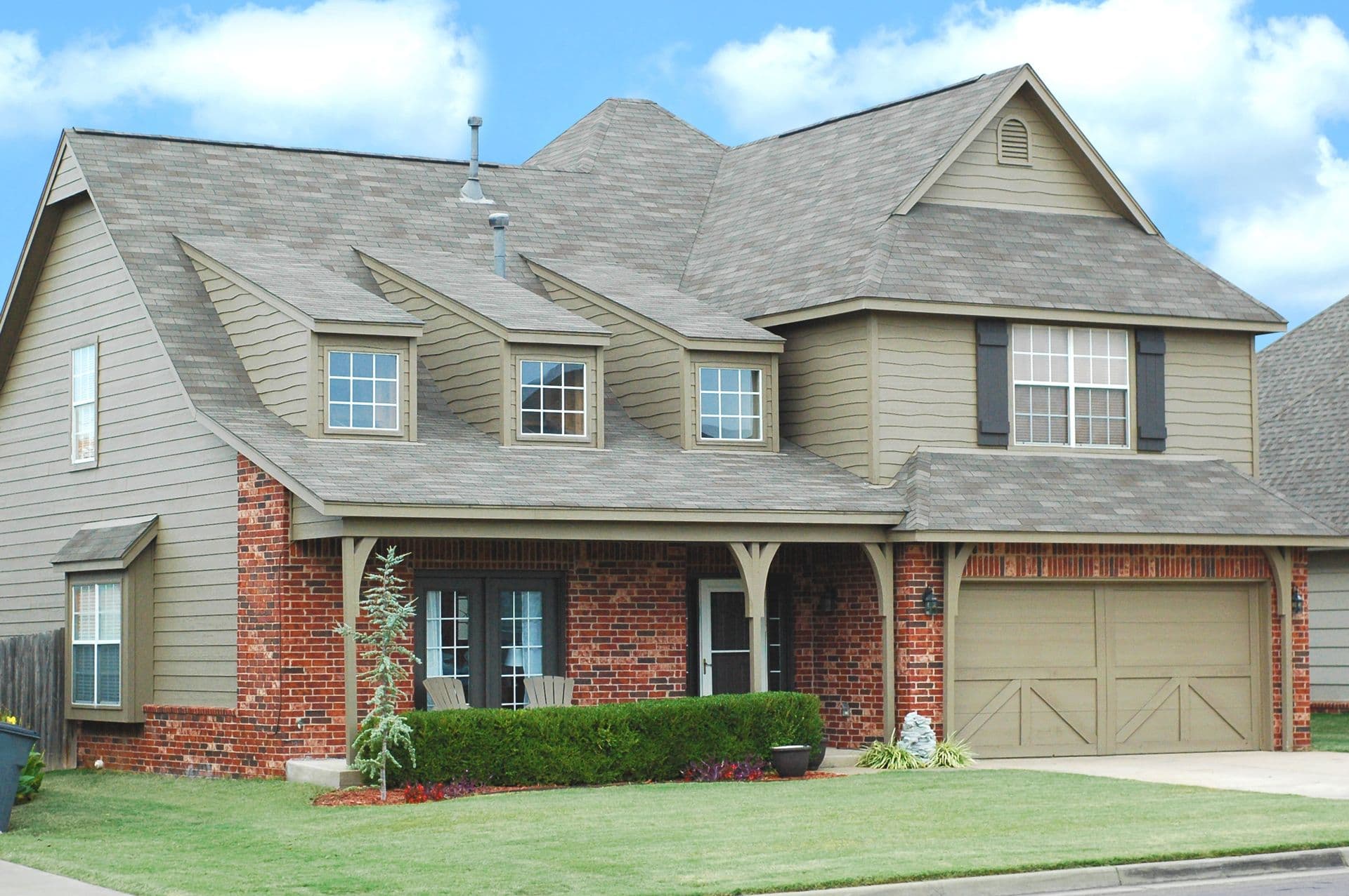 Two-story residential home with brick and siding exterior, multiple roof peaks, front porch, two-car garage, and landscaped lawn with shrubs and flowers