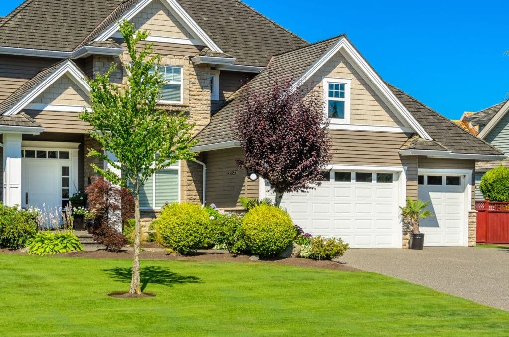 Two-story residential home with tan and brick exterior, white garage doors, manicured front lawn with young trees and landscaped shrubs under clear blue sky