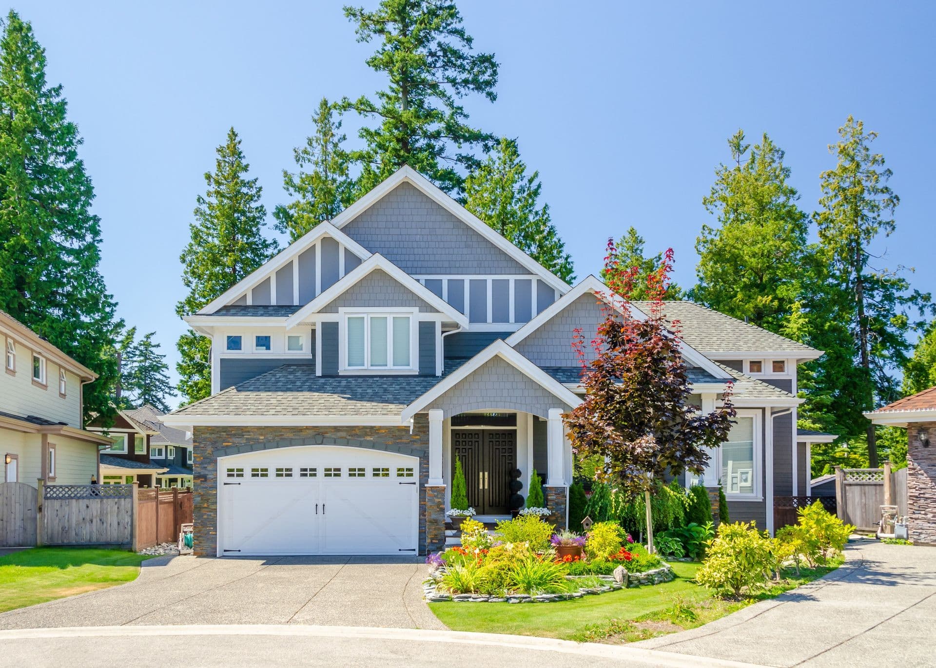 Modern two-story home with blue and white exterior, stone accents, white garage door, black front door, landscaped front yard with colorful plants, surrounded by tall evergreen trees