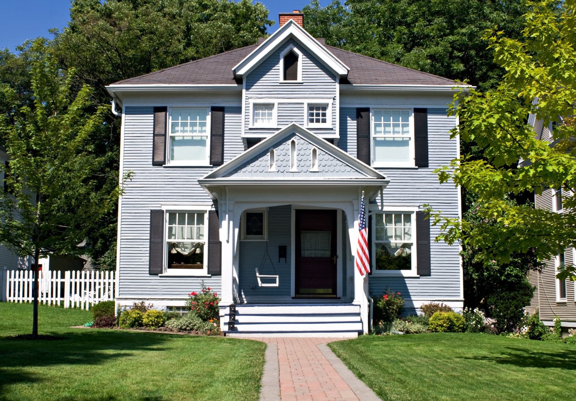 Two-story white Colonial house with dark shutters, brick chimney, and American flag on front porch, surrounded by mature trees and white picket fence