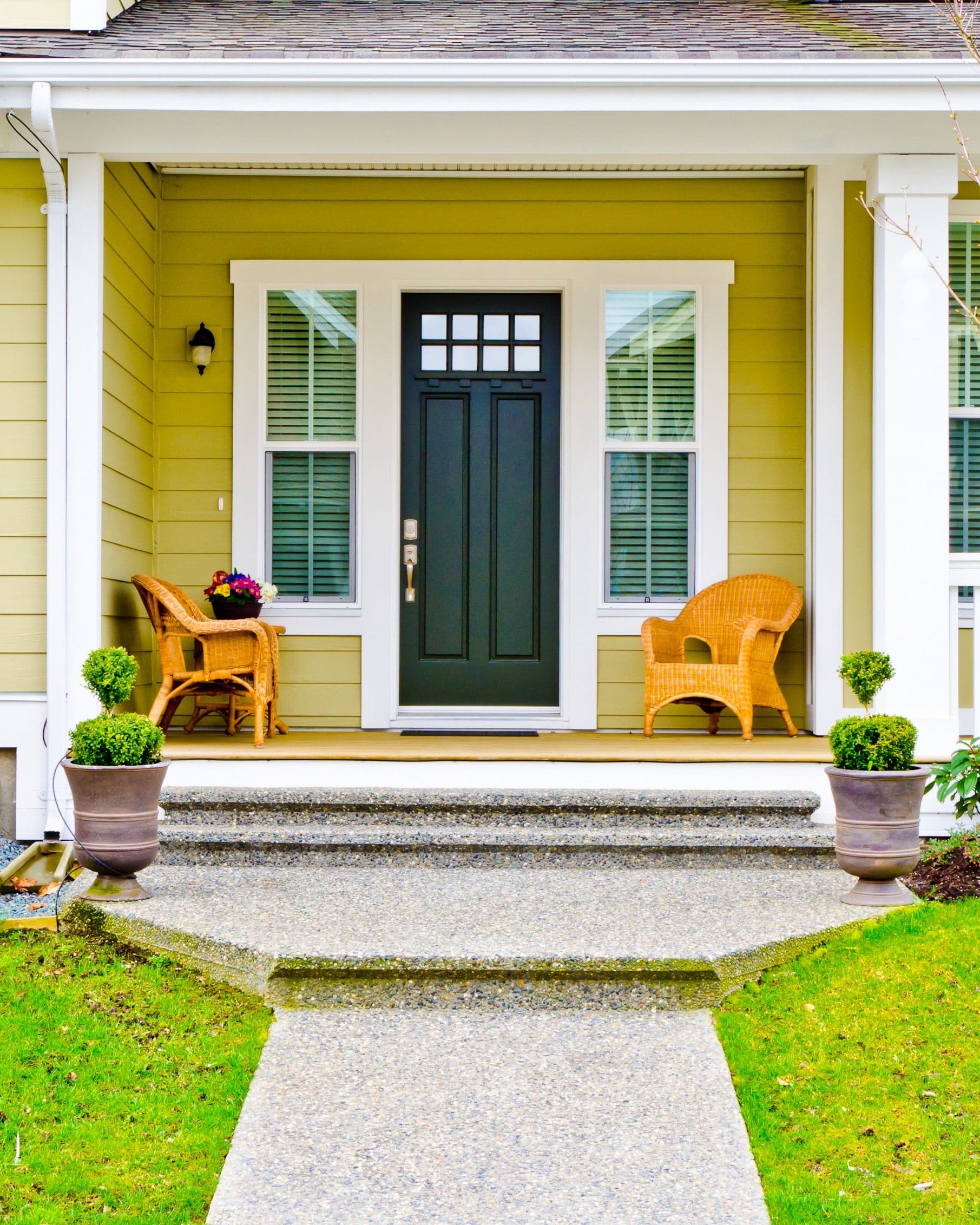Yellow house front porch with dark green door, white trim, wicker chairs, potted plants, and shuttered windows flanking the entry