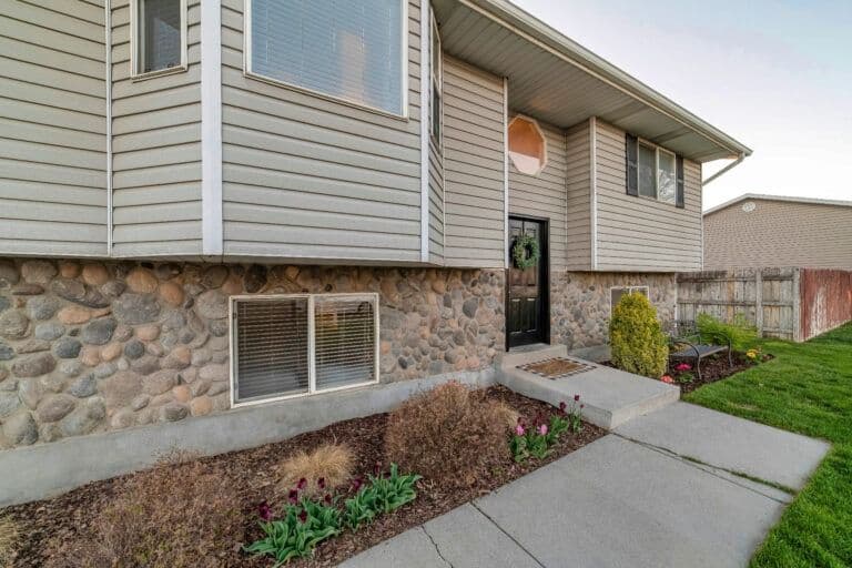 Single-story residential home with stone foundation, tan siding, and covered entryway, landscaped front yard with mulch beds and shrubs