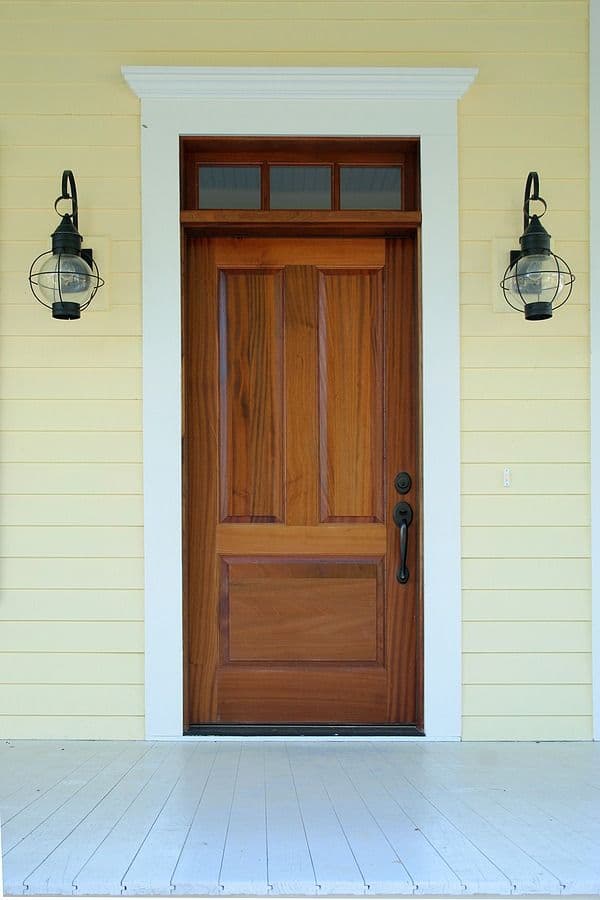 Front entrance with wooden door, transom window, white trim, and matching lantern sconces on pale yellow siding above white porch floor